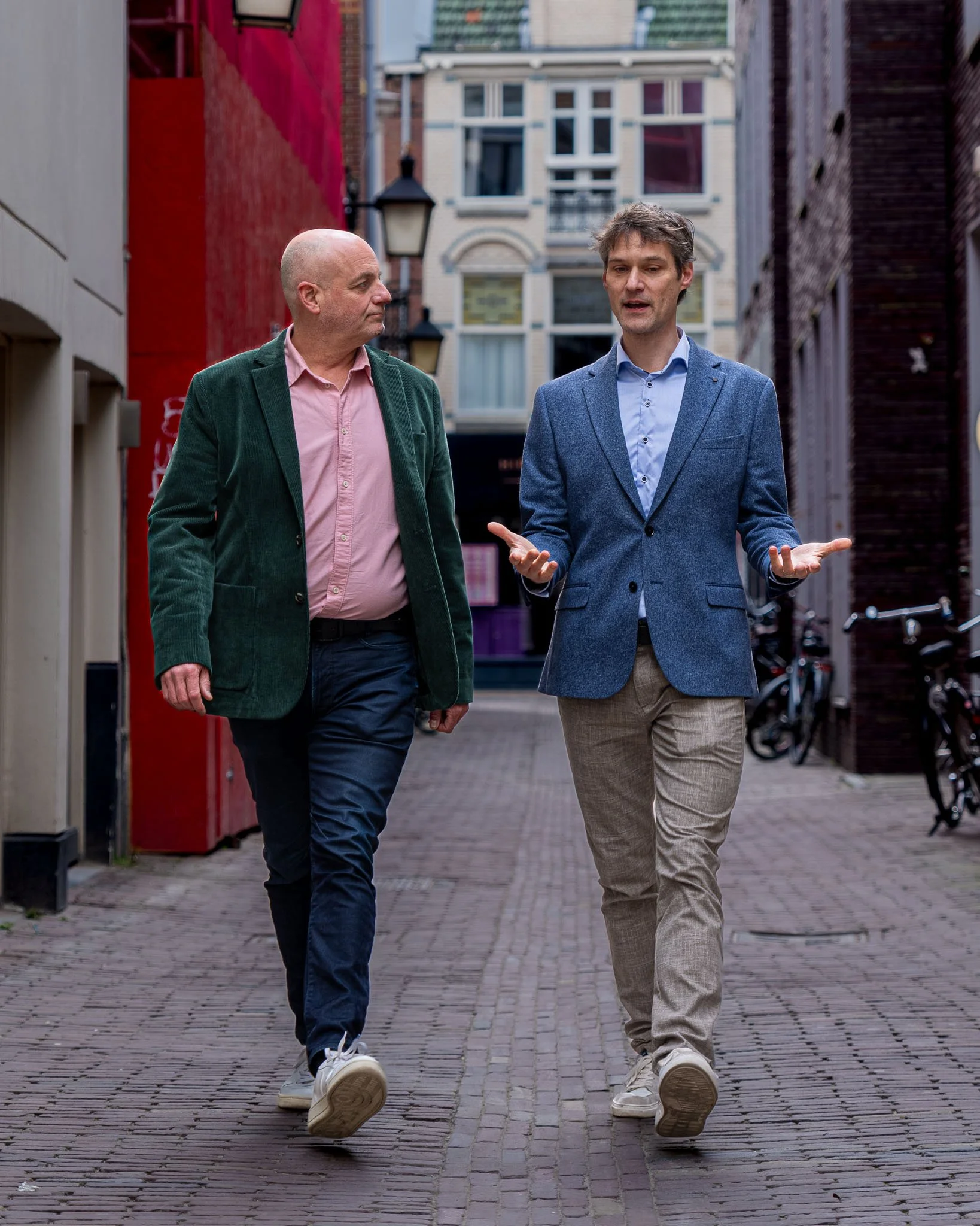 Twee mannen die samen wandelen in een stad, onderling in gesprek, omgeven door kleurrijke gebouwen en fietsen.