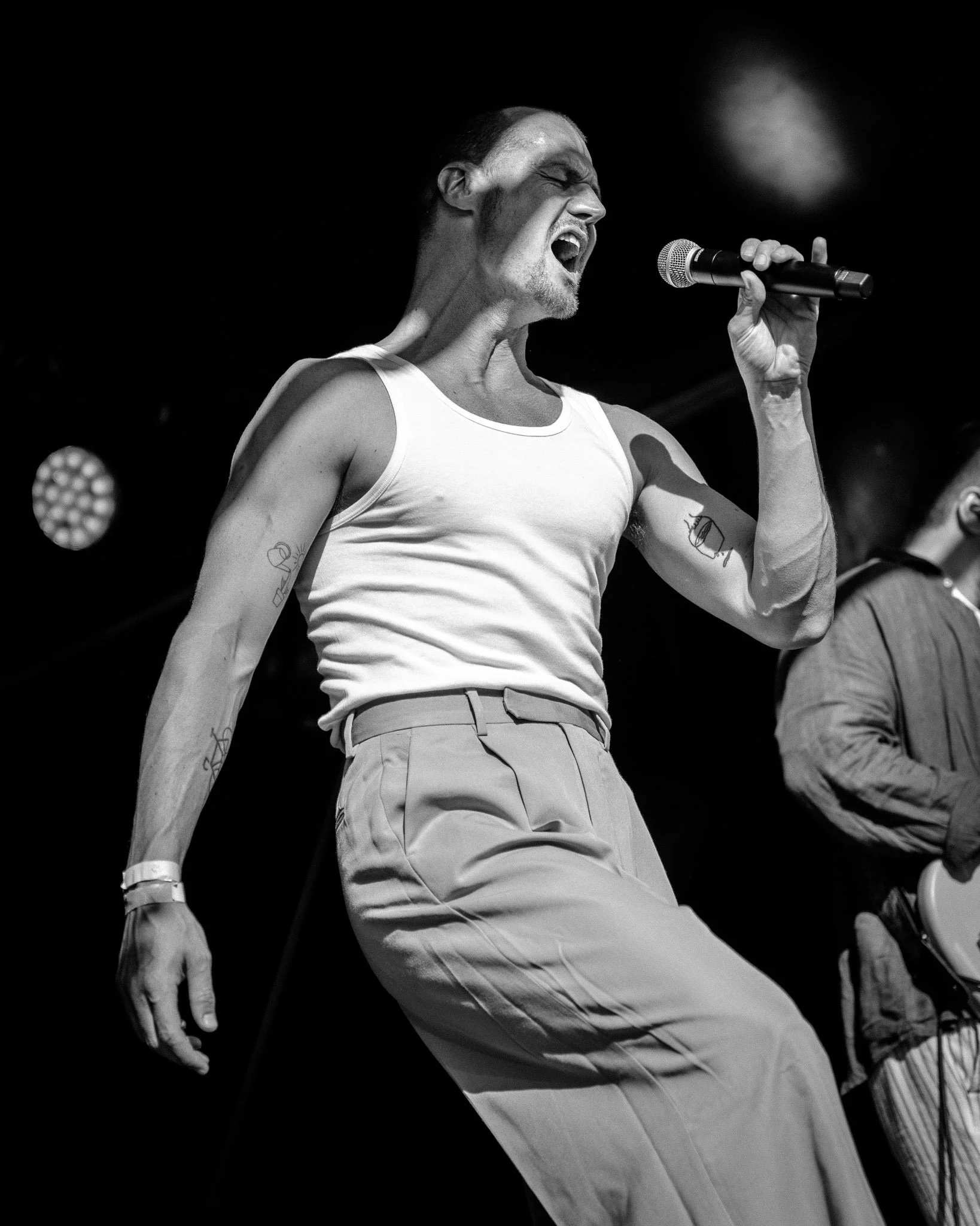 Zwart-wit foto van een man die schreeuwt terwijl hij een microfoon vasthoudt op een podium, gekleed in een witte tanktop en lichtgekleurde broeken.