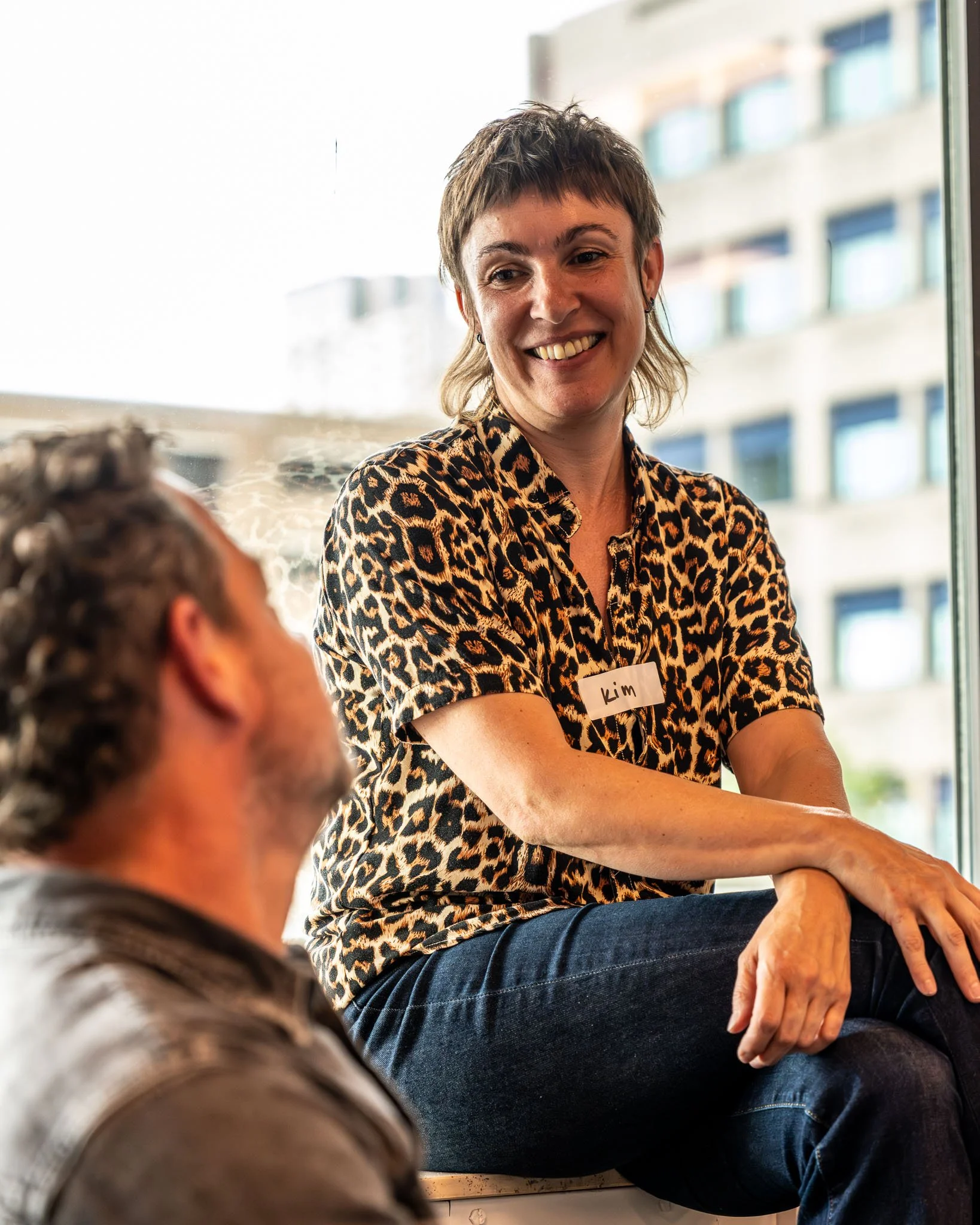Een vrouw met kort bruin haar, lachend en een leopardprint blouse, zit op een stoel en heeft een naamkaartje met de naam Kim. Ze zit binnen bij een raam met uitzicht op een appartementencomplex.