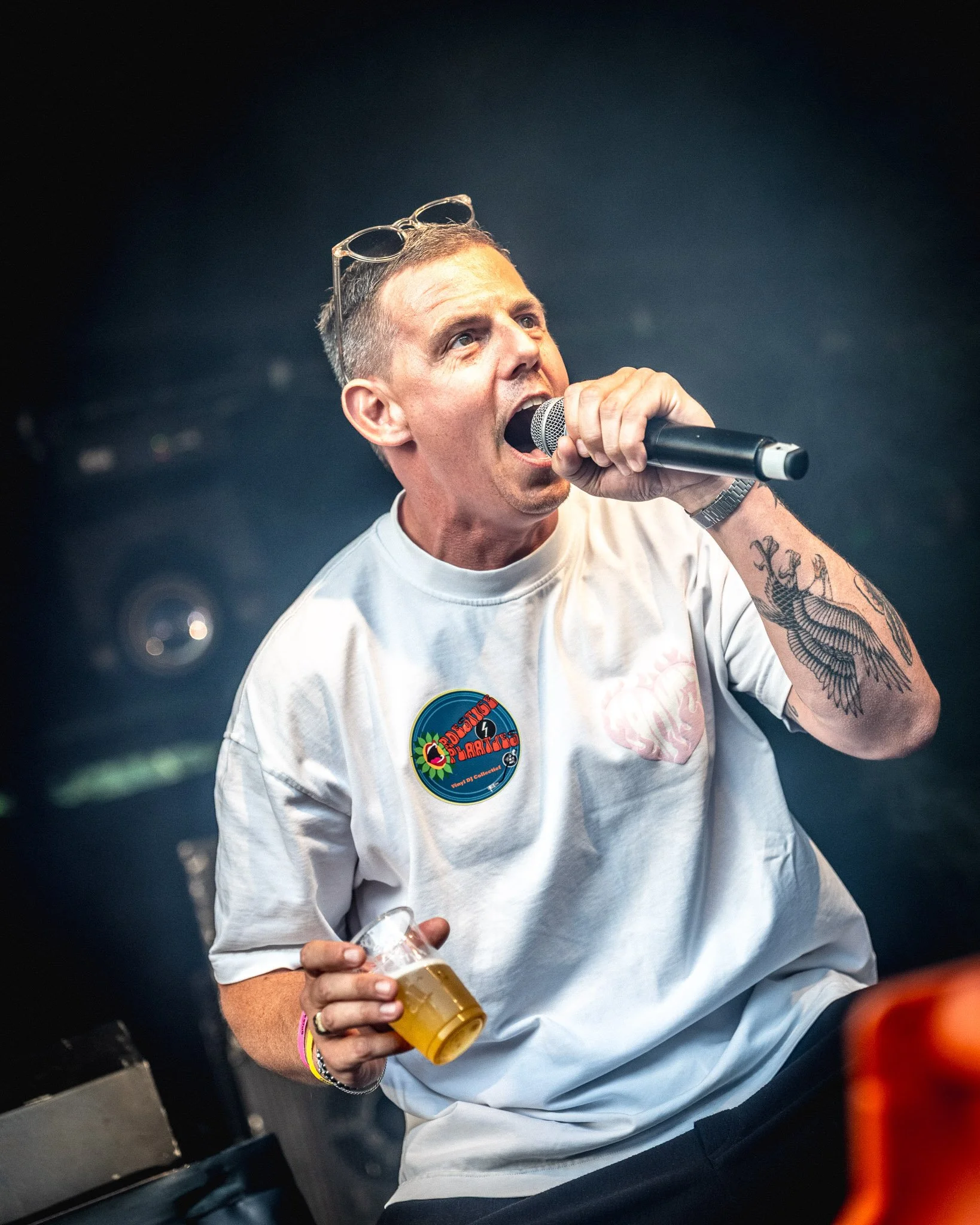 Man met kort haar en tatoeages op zijn arm drinkt uit een glas met een drankje, hij draagt een witte T-shirt met een sticker.