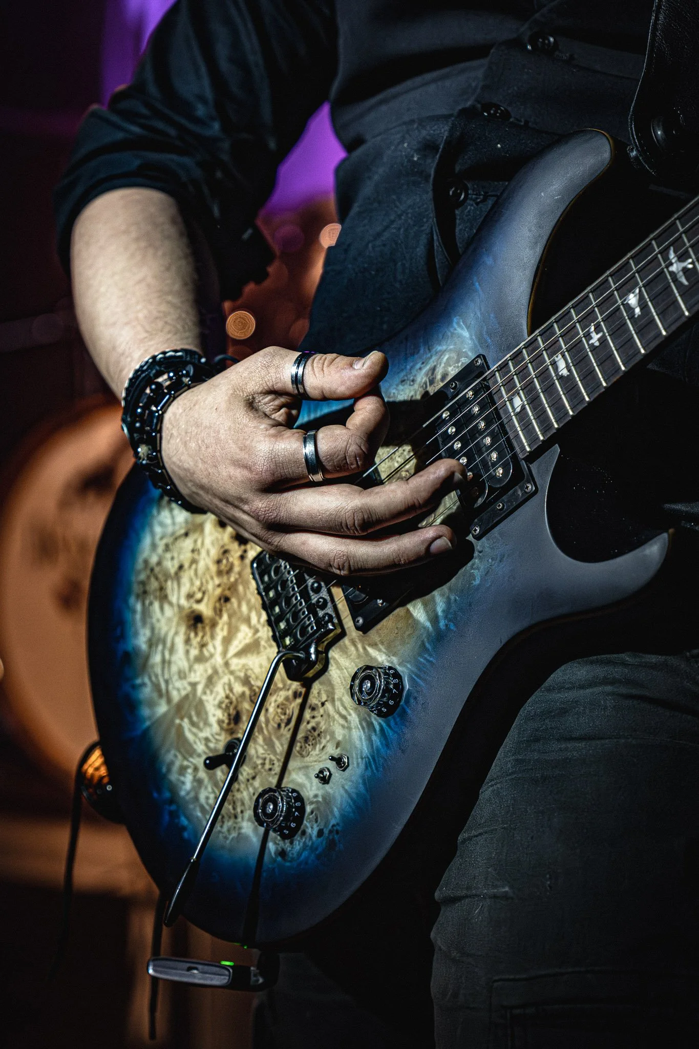 Persoon speelt elektrische gitaar, draag horloge en meerdere armbanden, achtergrond donker met kleurrijke lichtvlekken.