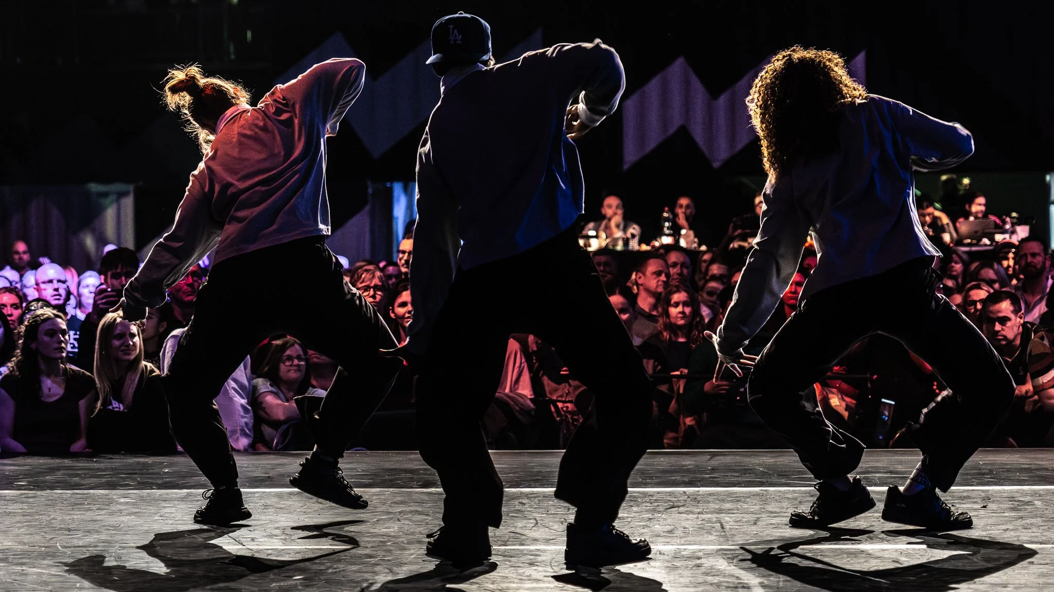 Drie dansers in zwarte broeken en donkere jassen dansen op een podium, met een publiek dat toekijkt op de achtergrond.
