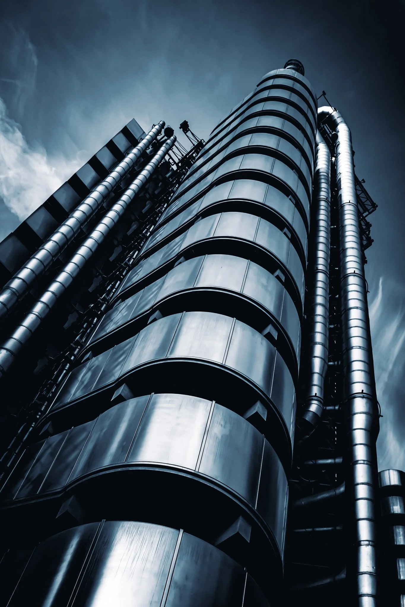 Een lager perspectief van een modern, hoog gebouw met een metalen buitenkant, pijpen en ventilatie- en onderhoudsstructuren zichtbaar aan de zijkant, onder een bewolkte hemel.