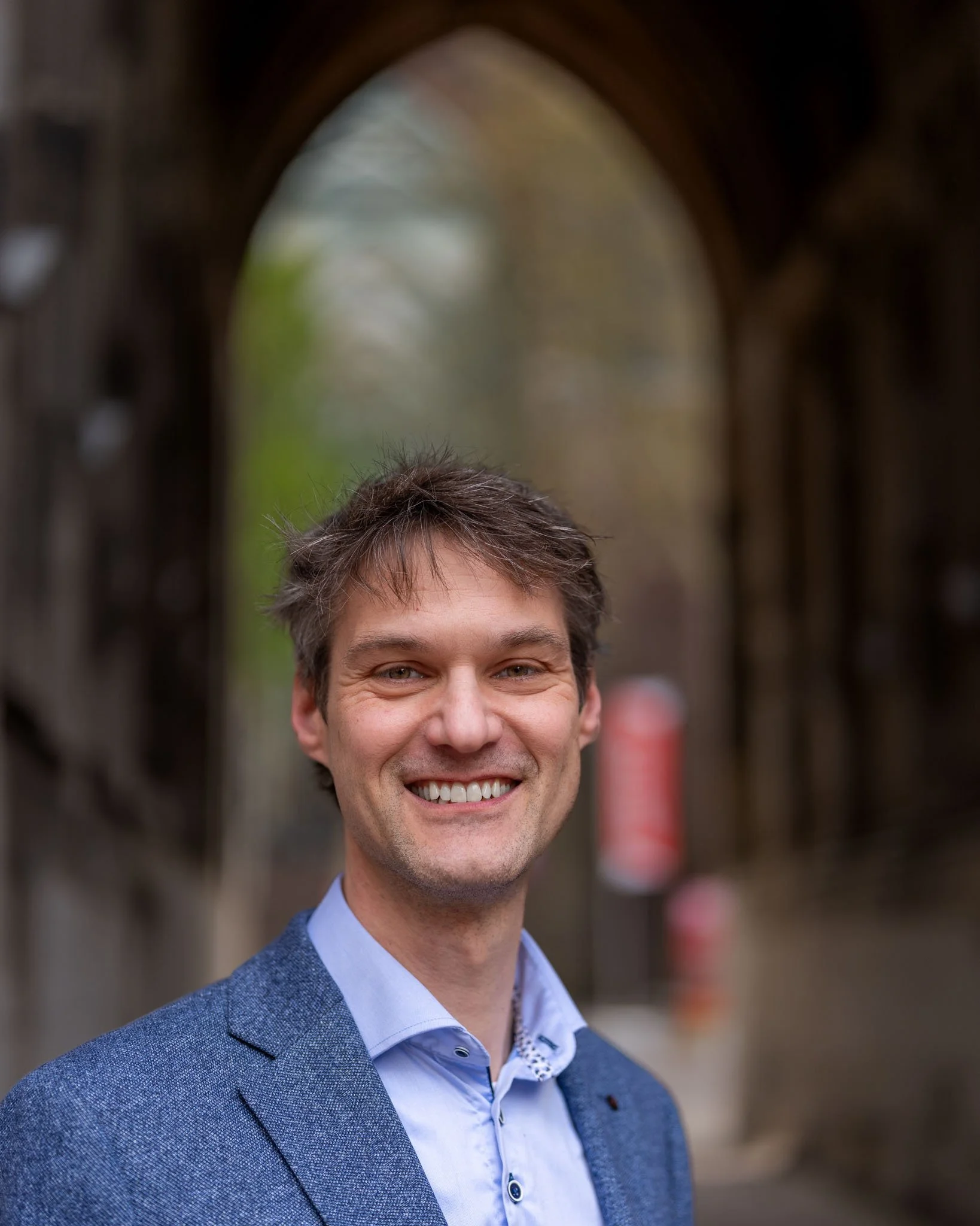 Portret van een lachende man met bruin haar en lichte huid, gekleed in een blauwe blazer en een lichtblauw overhemd, buiten met een kerkachtig gebouw op de achtergrond.