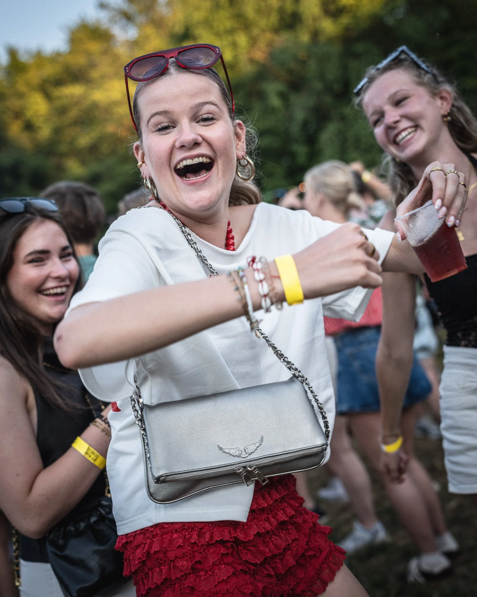 Lachende jonge vrouwen op een festival, een vrouw draagt een witte blouse, rode korte rok en een witte tas met een zilveren klok. Ze heeft ook rode sieraden en draagt een bril op haar hoofd. De achtergrond laat een ondergaande zon en groene bomen zie