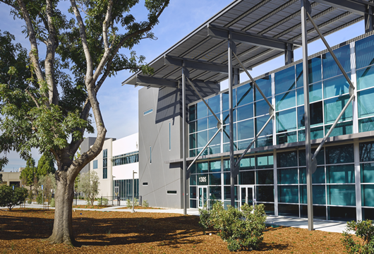Modern office building with glass facade and metal framework, surrounded by landscaped area with trees and bushes.