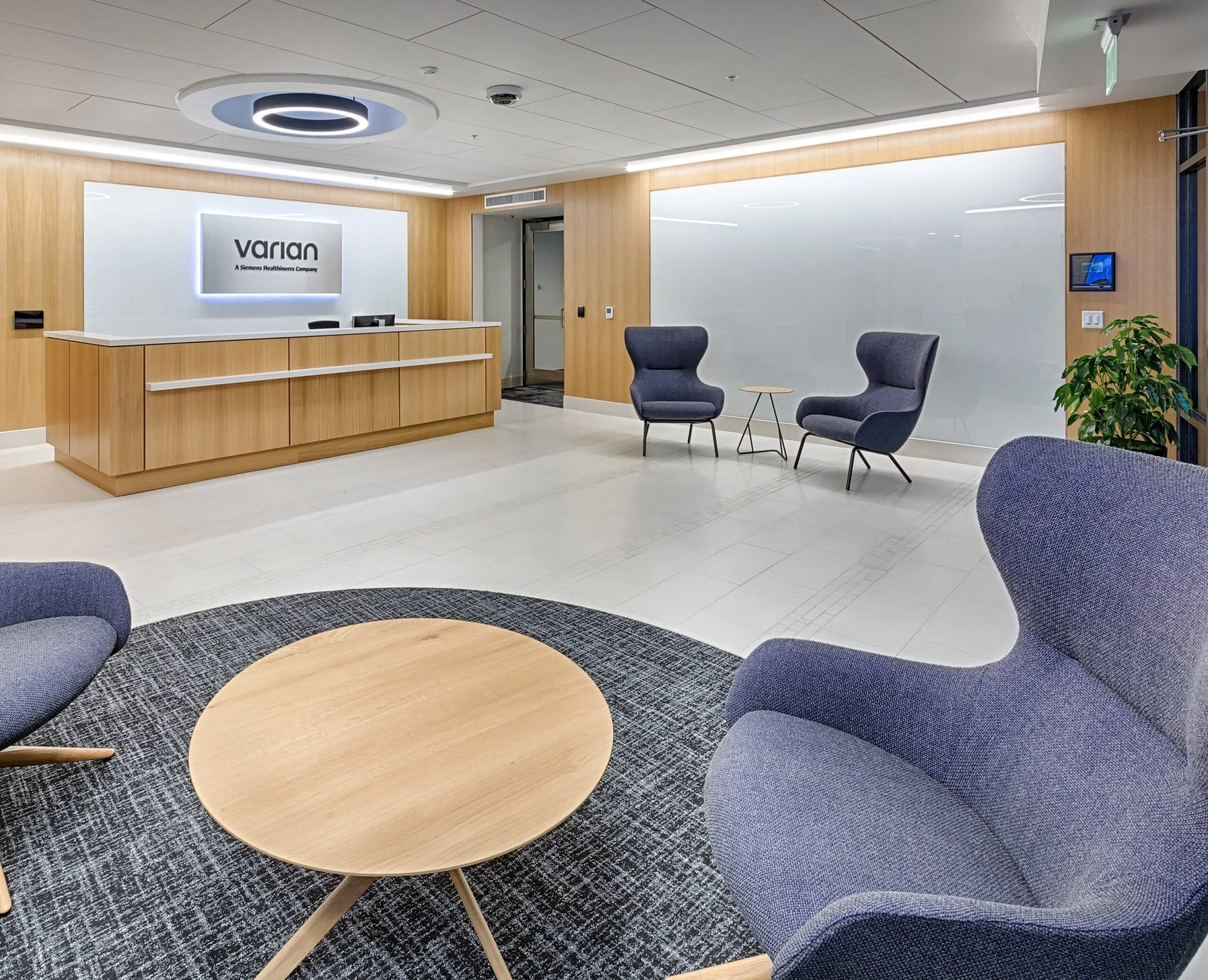 Modern office reception area with a wooden front desk, a white logo sign reading 'Varian,' comfortable gray armchairs, a small round wooden table, a large window, indoor plants, and a digital screen on the wall.
