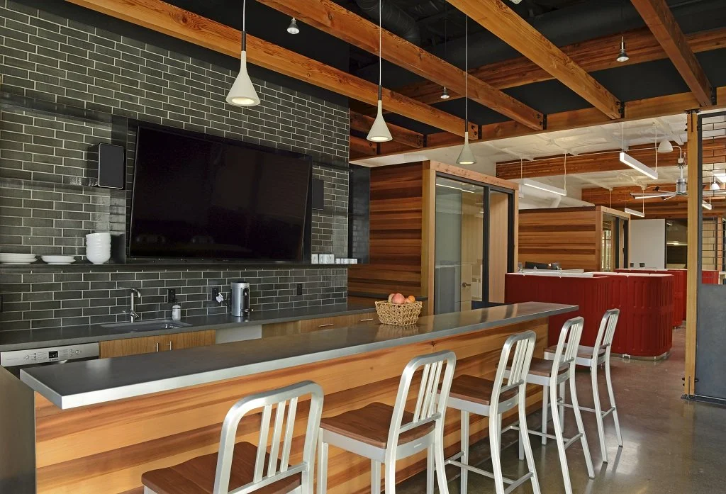 Modern kitchen with wooden beams on ceiling, black brick wall, flat-screen TV, and a long counter with four metal chairs.