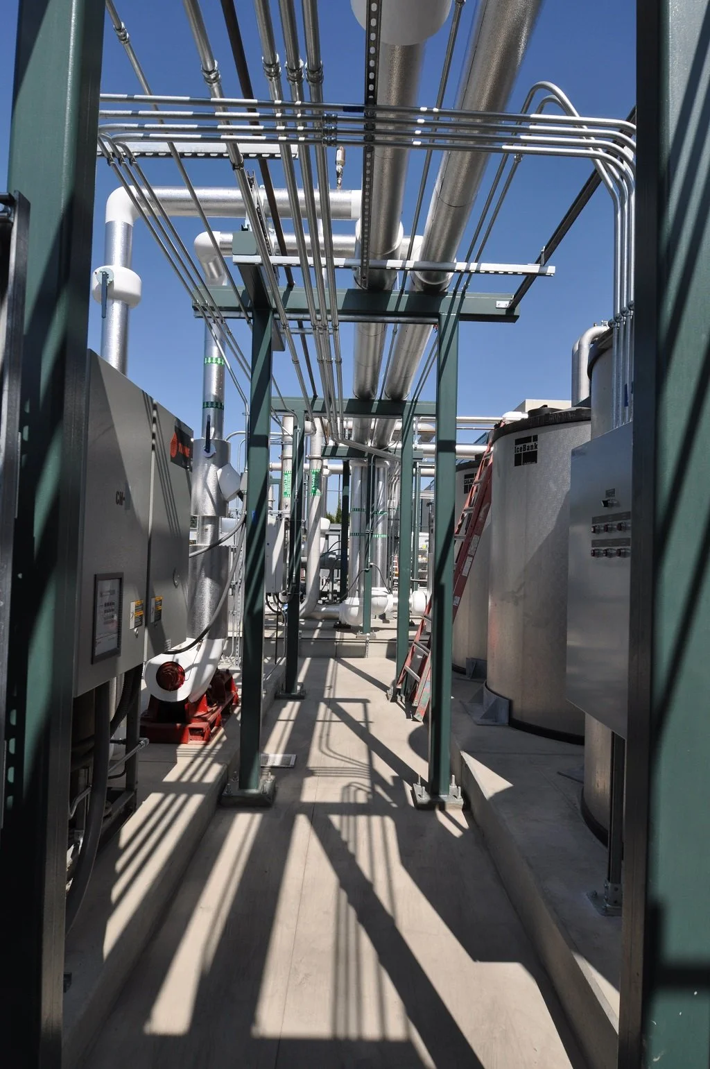 Industrial site with pipes, electrical panels, and metal framework on a concrete floor under a clear blue sky.