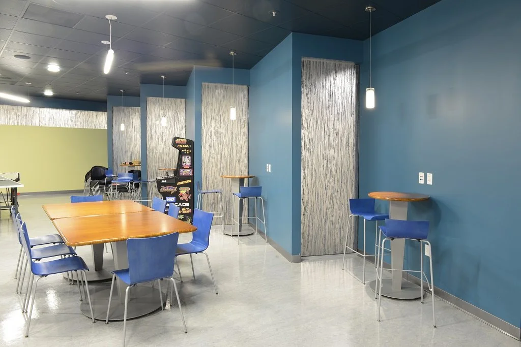 Modern indoor seating area with a wooden table surrounded by blue chairs, small high-top tables with blue chairs along a colorful blue wall, pendant lights, and arcade machines in the background.