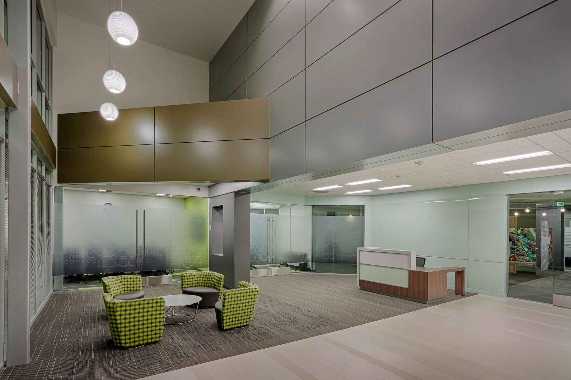 Modern office lobby with a seating area of four green patterned armchairs around a small white table, glass doors and walls, a reception desk, and overhead lighting.