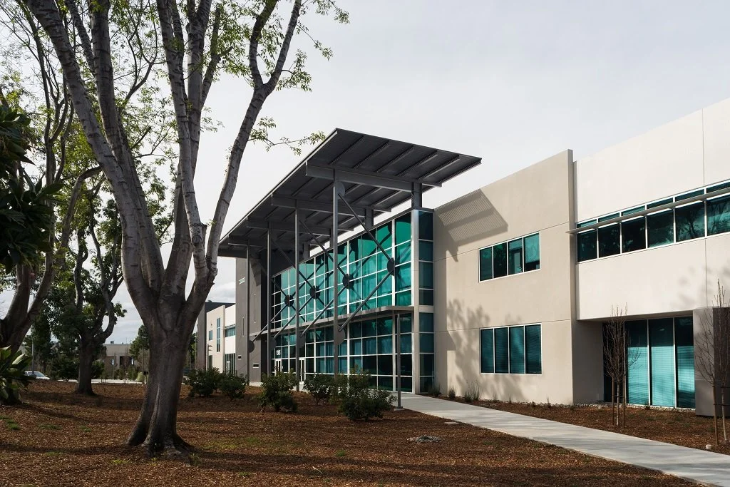 Modern office building with glass windows and solar panels on the roof, surrounded by trees and landscaped grounds.