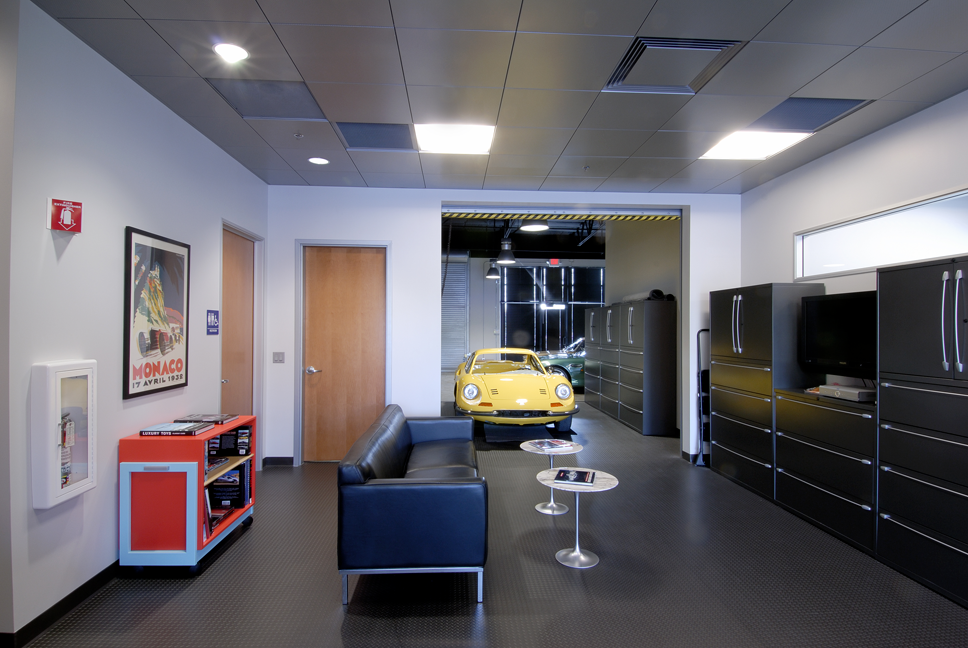 An office lounge area with a yellow vintage car parked inside. The room has a black couch, small tables, a TV, black storage cabinets, and a framed poster on the wall. There are two doors, windows on the right, and ceiling lights.