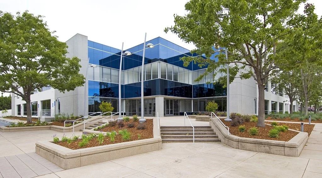 Modern building with glass windows, surrounded by trees and landscaped with plants and flower beds, with stairs leading to the entrance.