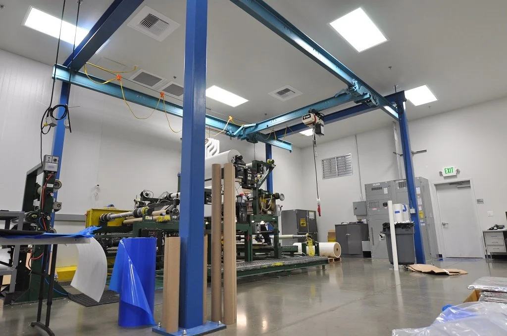 Industrial manufacturing room with machinery, blue support beams, and various equipment wrapped in blue and brown paper.