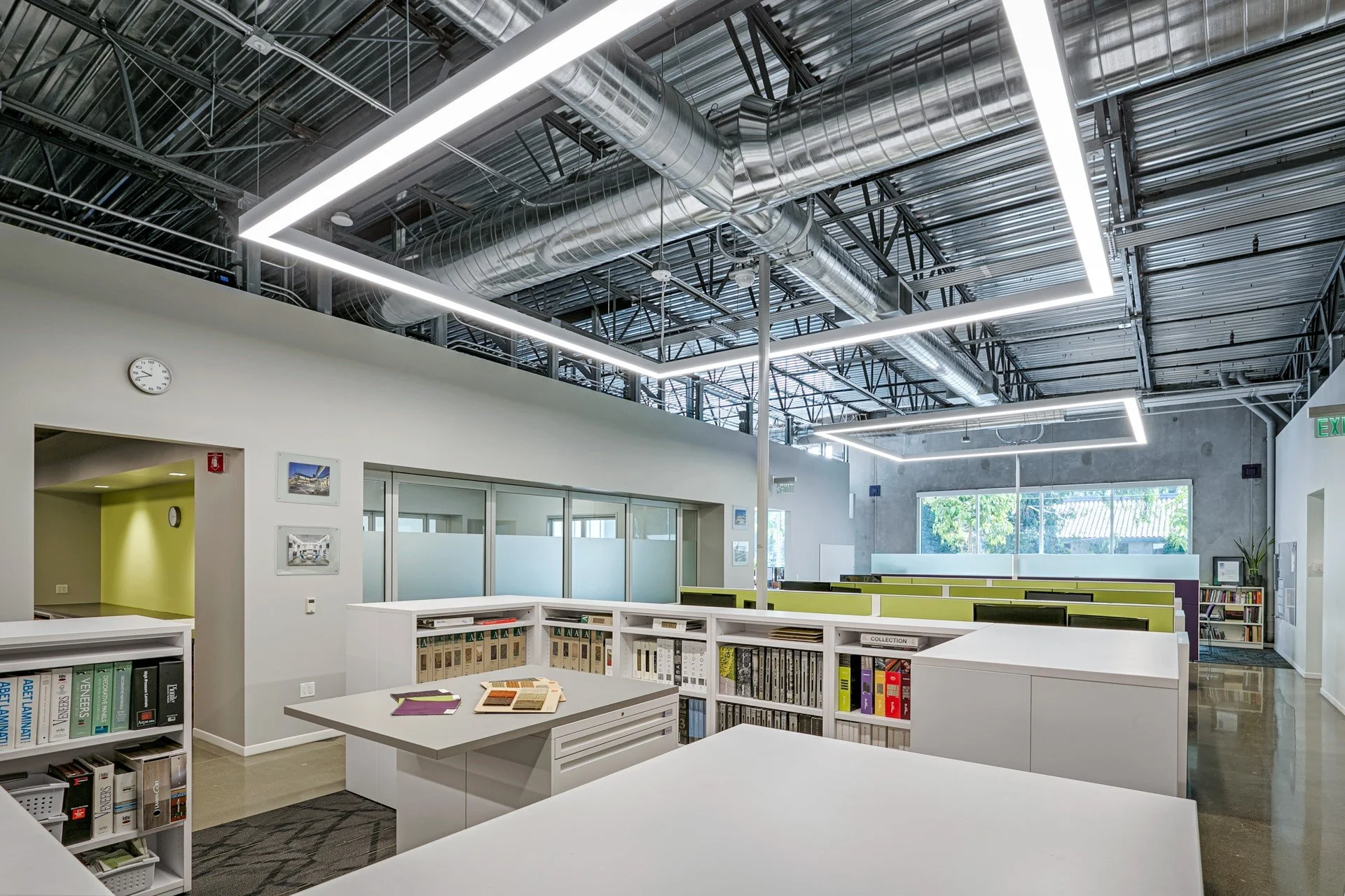 Modern office space with white desks, bookshelves, and green dividers, featuring an industrial ceiling with exposed pipes and ductwork, large window, and bright lighting.