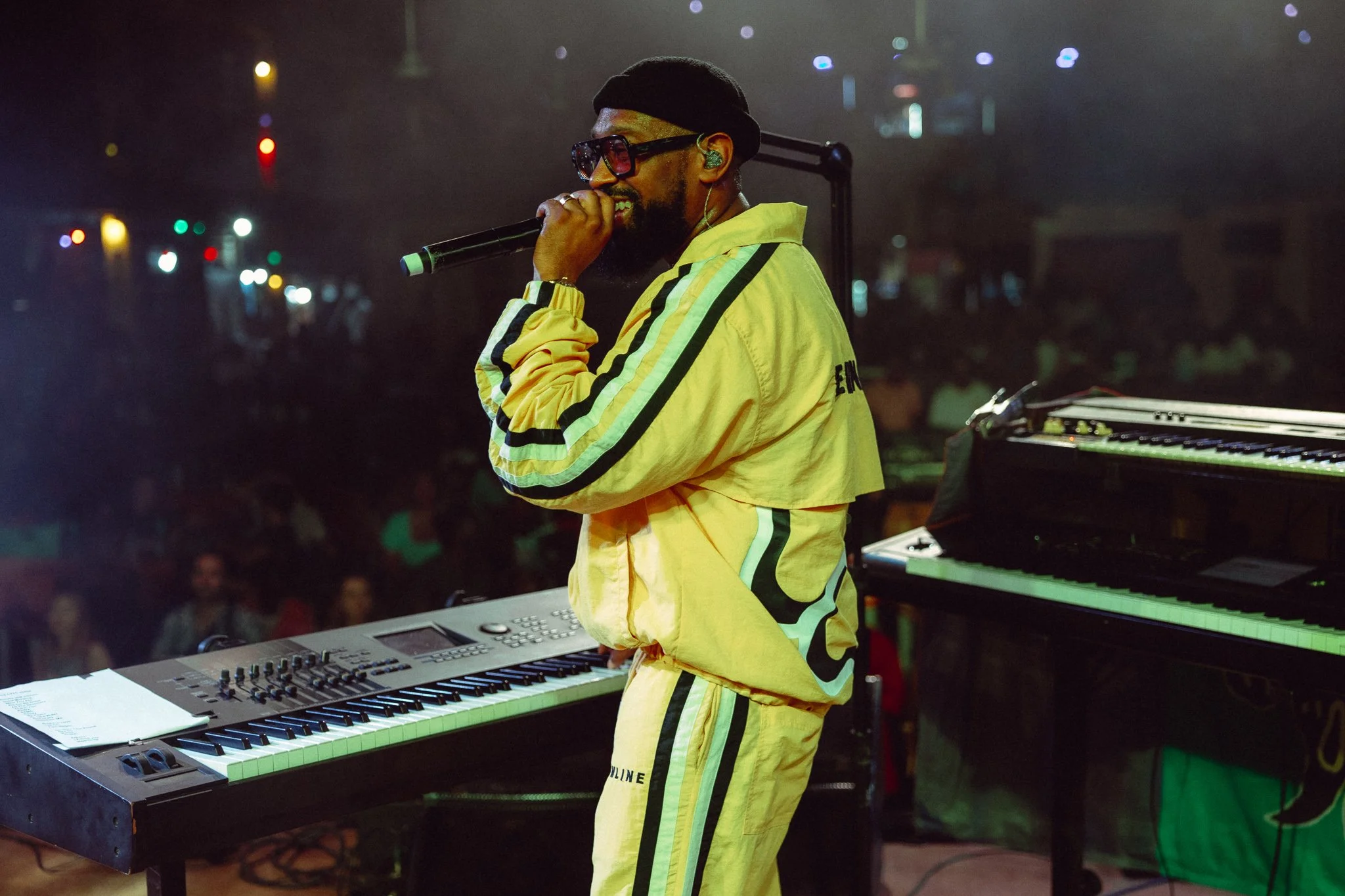 PJ Morton in yellow track suite with a black beanie on his head, holds a microphone with a smile on his face as he stands in front of two pianos.