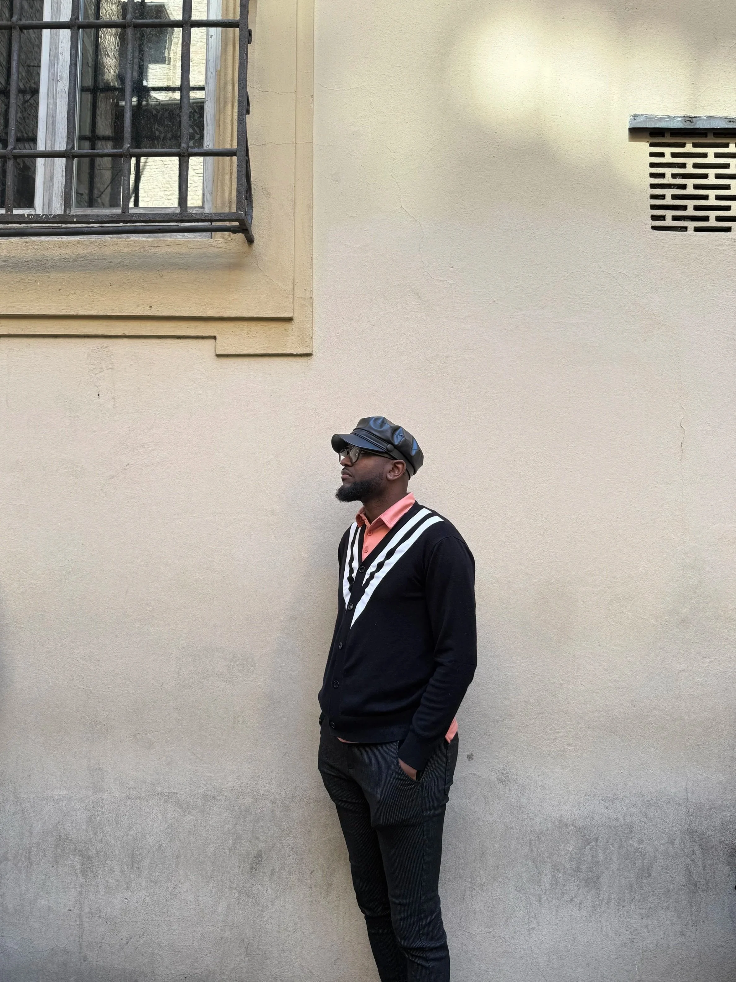 A man with a black leather hat, a salmon-colored shirt, black sweater with white trim on the collar, and dark colored jeans, stands in front of a cream-colored wall.