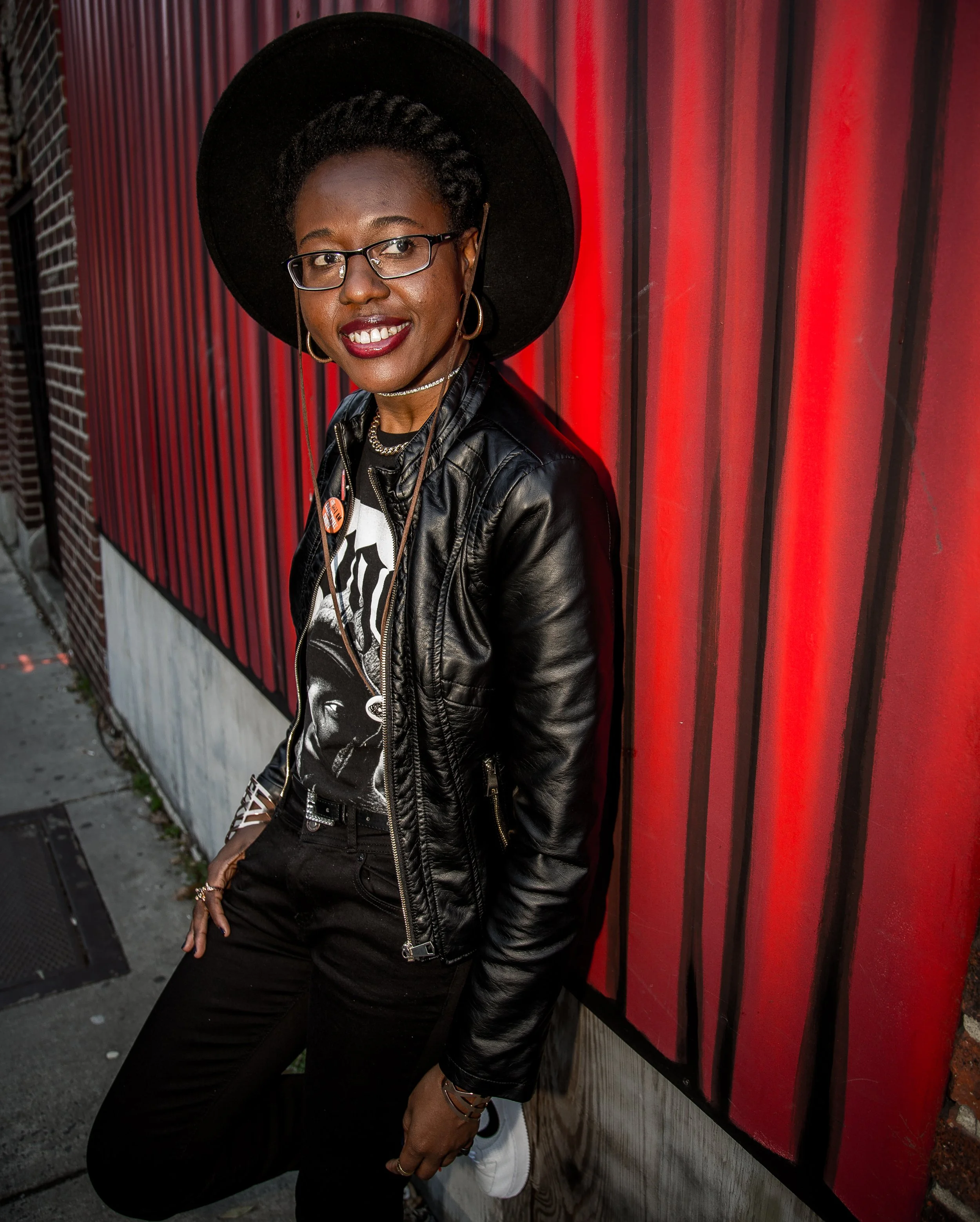 A woman (Kia O. Moore) in all black attire wears a large brimmed black hat and black leather coat. She leans up against a painted red wall. She smiles and glances off screen.