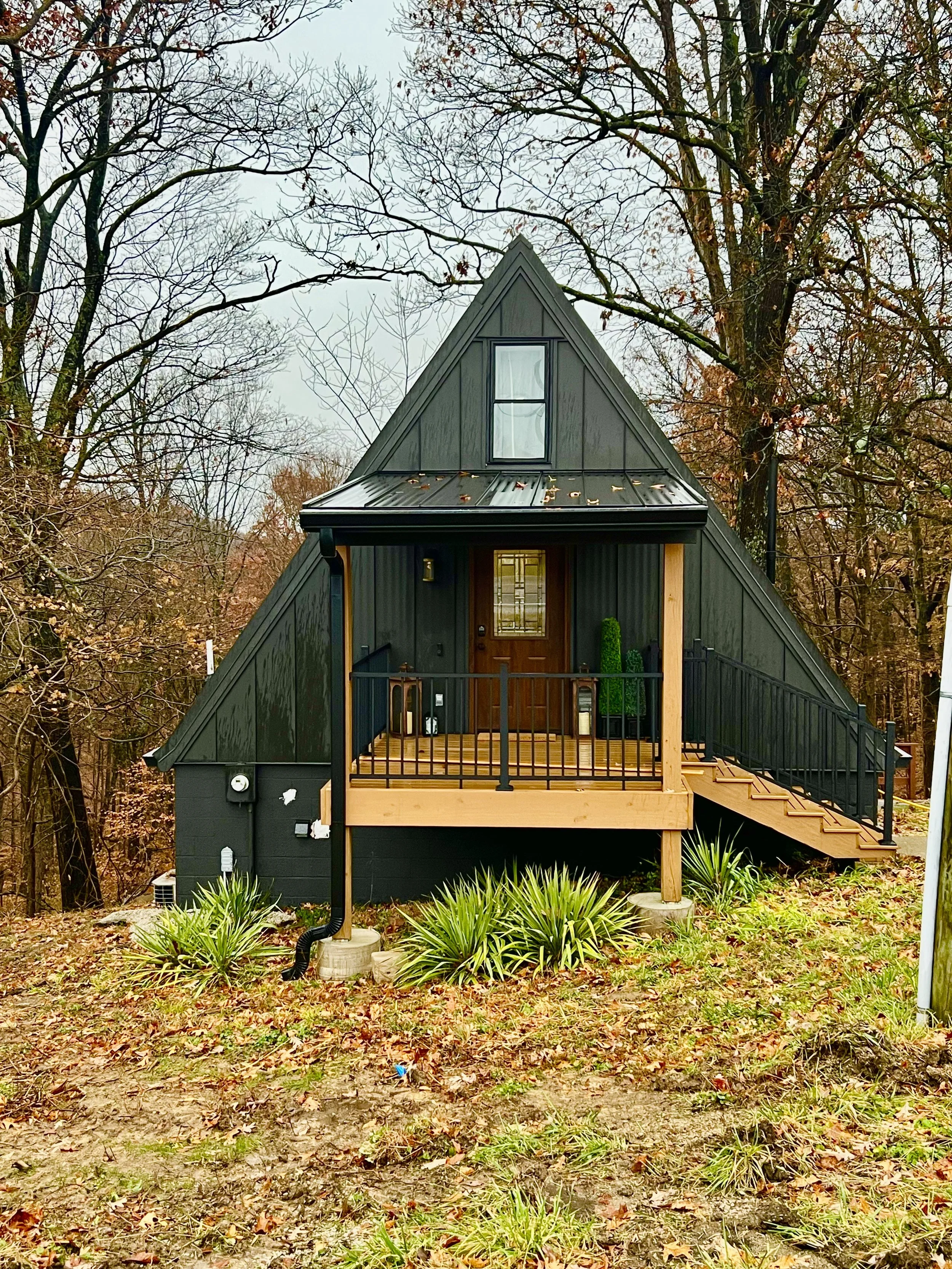 Small black house with a steep triangular roof, metal roofing, and a front porch with steps, located in a wooded area with trees and grass.