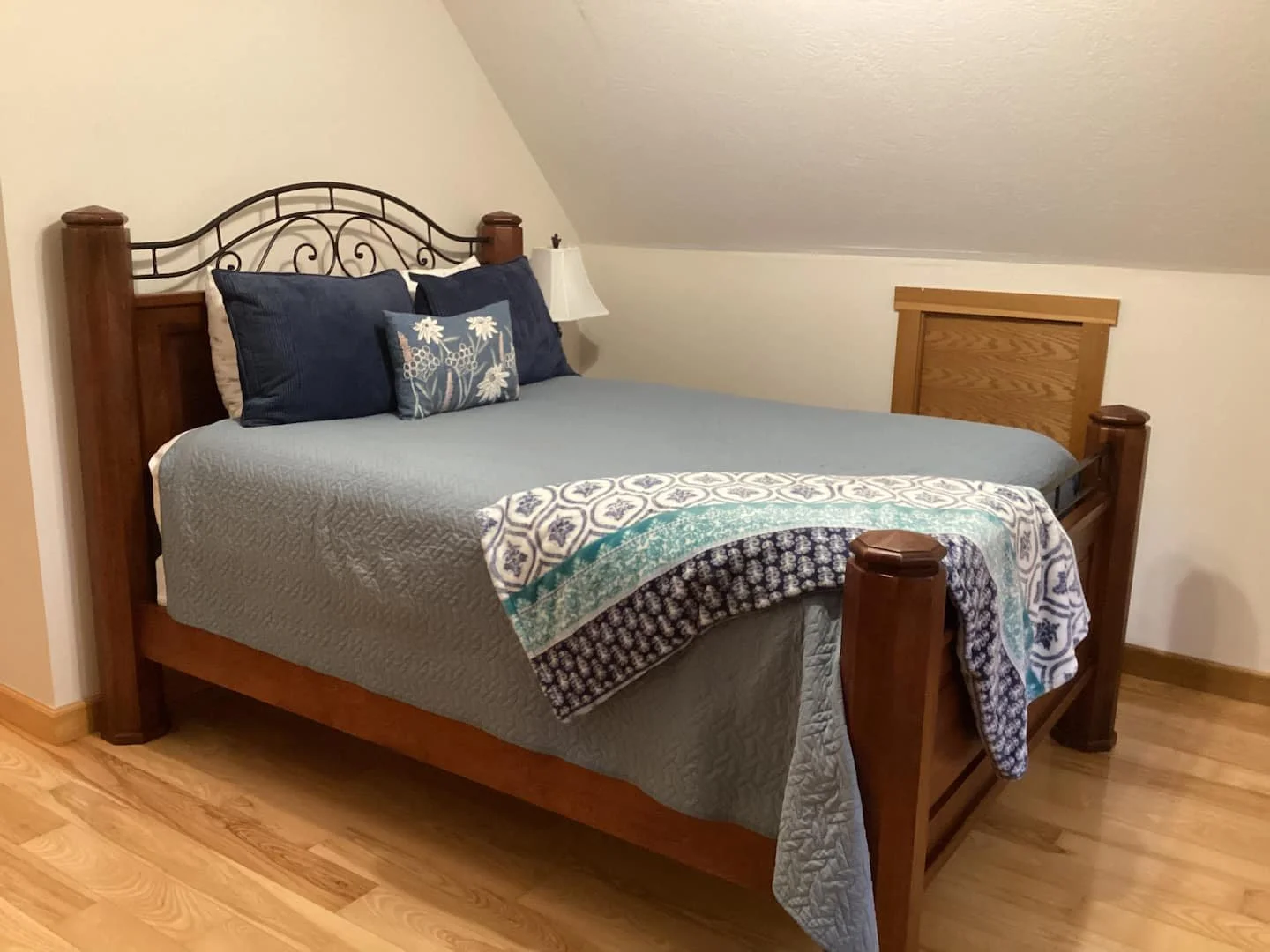A bedroom with a wooden bed frame, blue pillows, a floral pillow, a quilted bed cover, and a bedside lamp, located under a sloped ceiling.