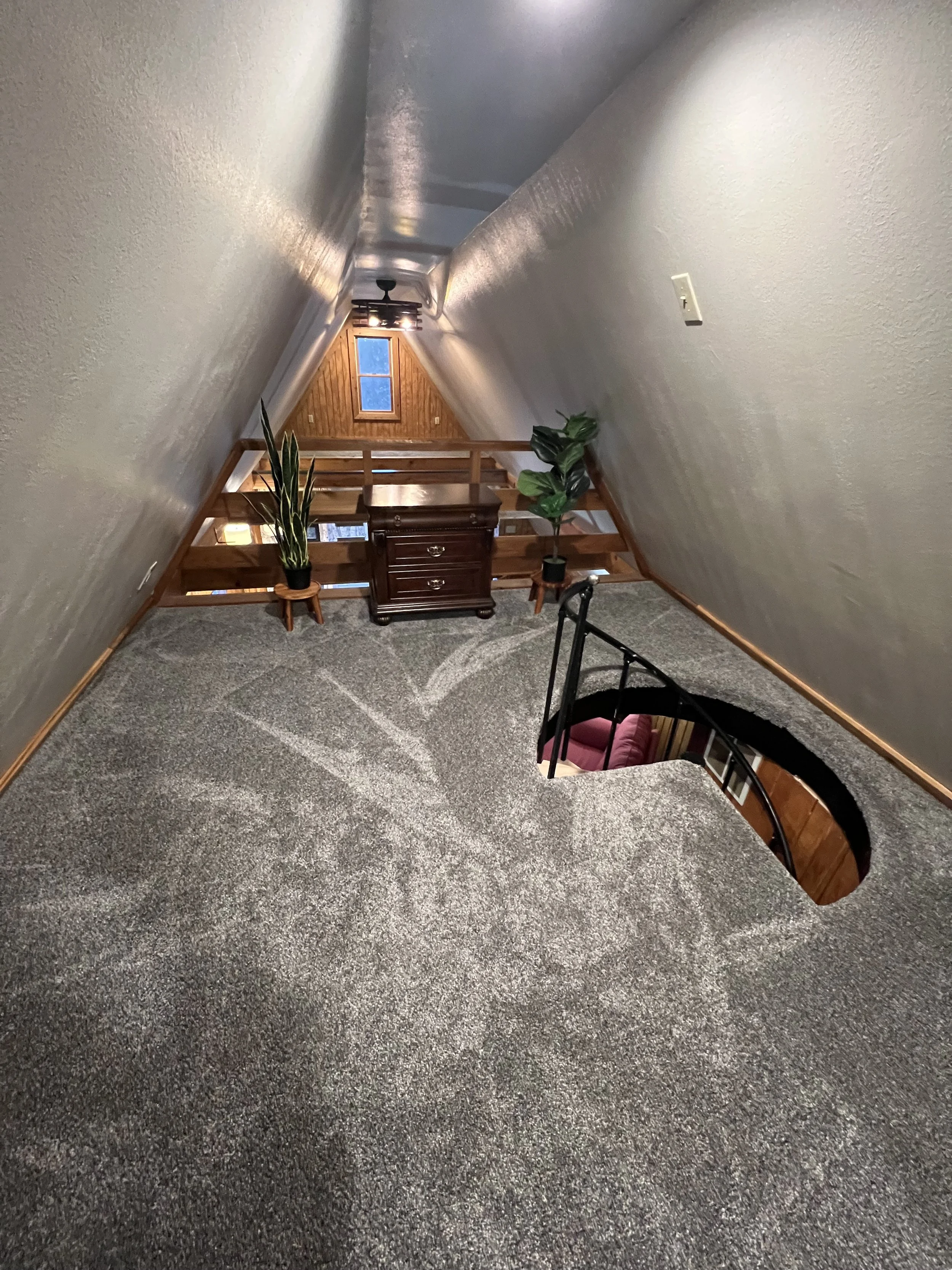 View of a cozy attic room with sloped ceiling, carpeted floor, wooden railing, and furniture including a dresser, potted plants, and a window with an outdoor view.