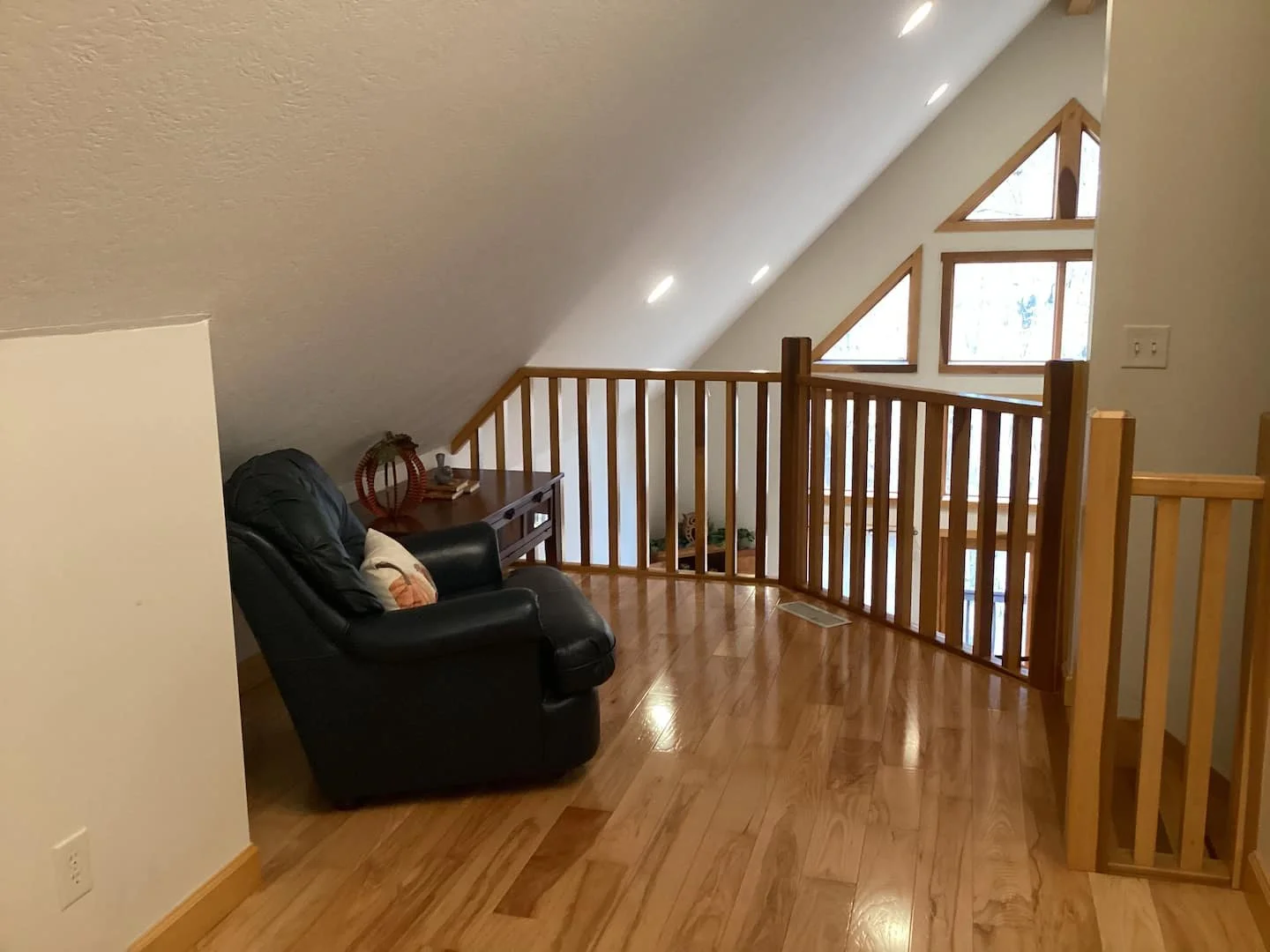 A cozy attic space with hardwood flooring, a black leather armchair, a small wooden side table with decorative items, a wooden railing, and triangular windows letting in natural light.