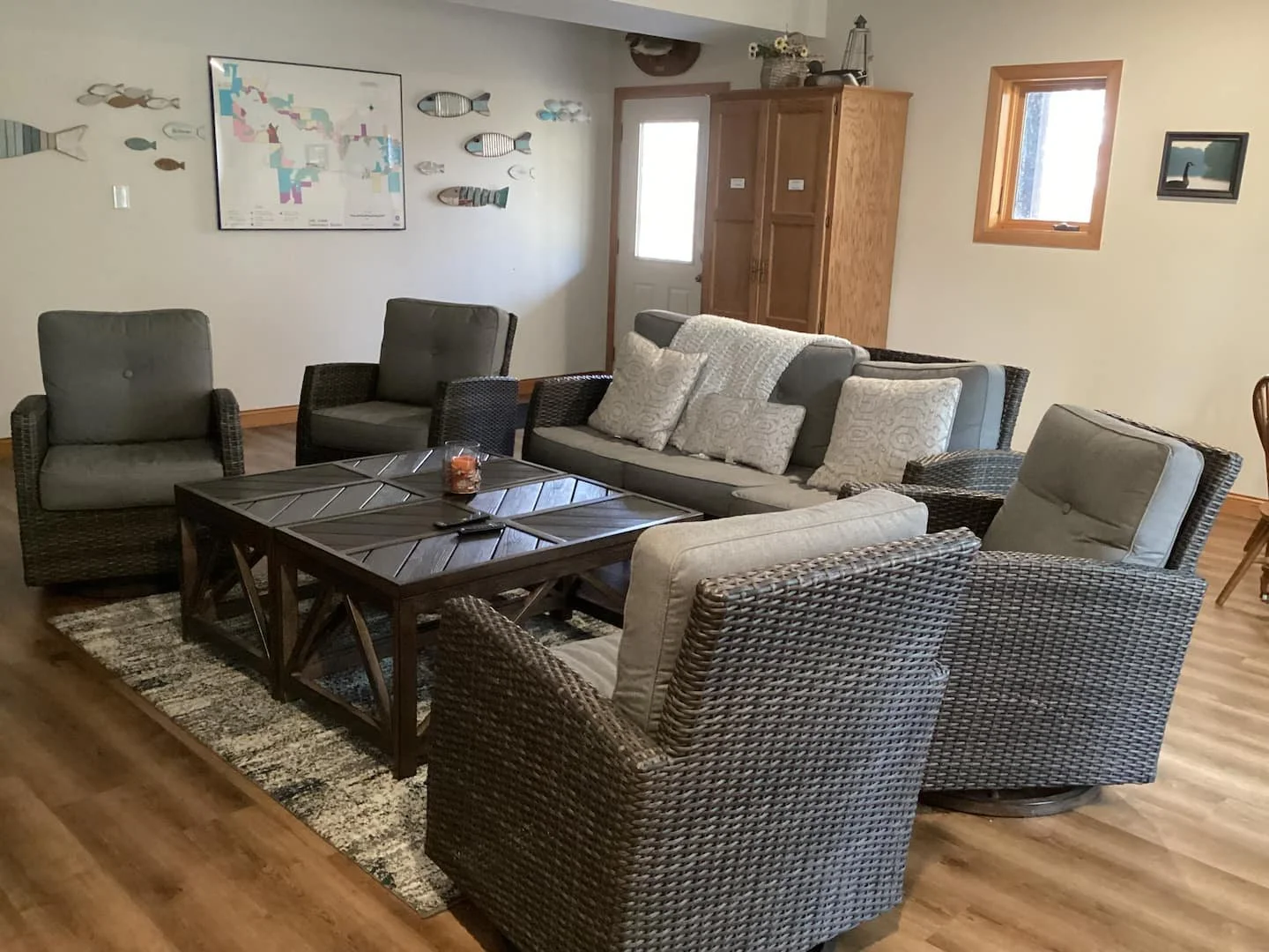 Living room with wicker and upholstered chairs around a wooden coffee table, beige sofa and pillows, wooden floor, wall art including a map and fish decorations, window, door, and wooden cabinet.