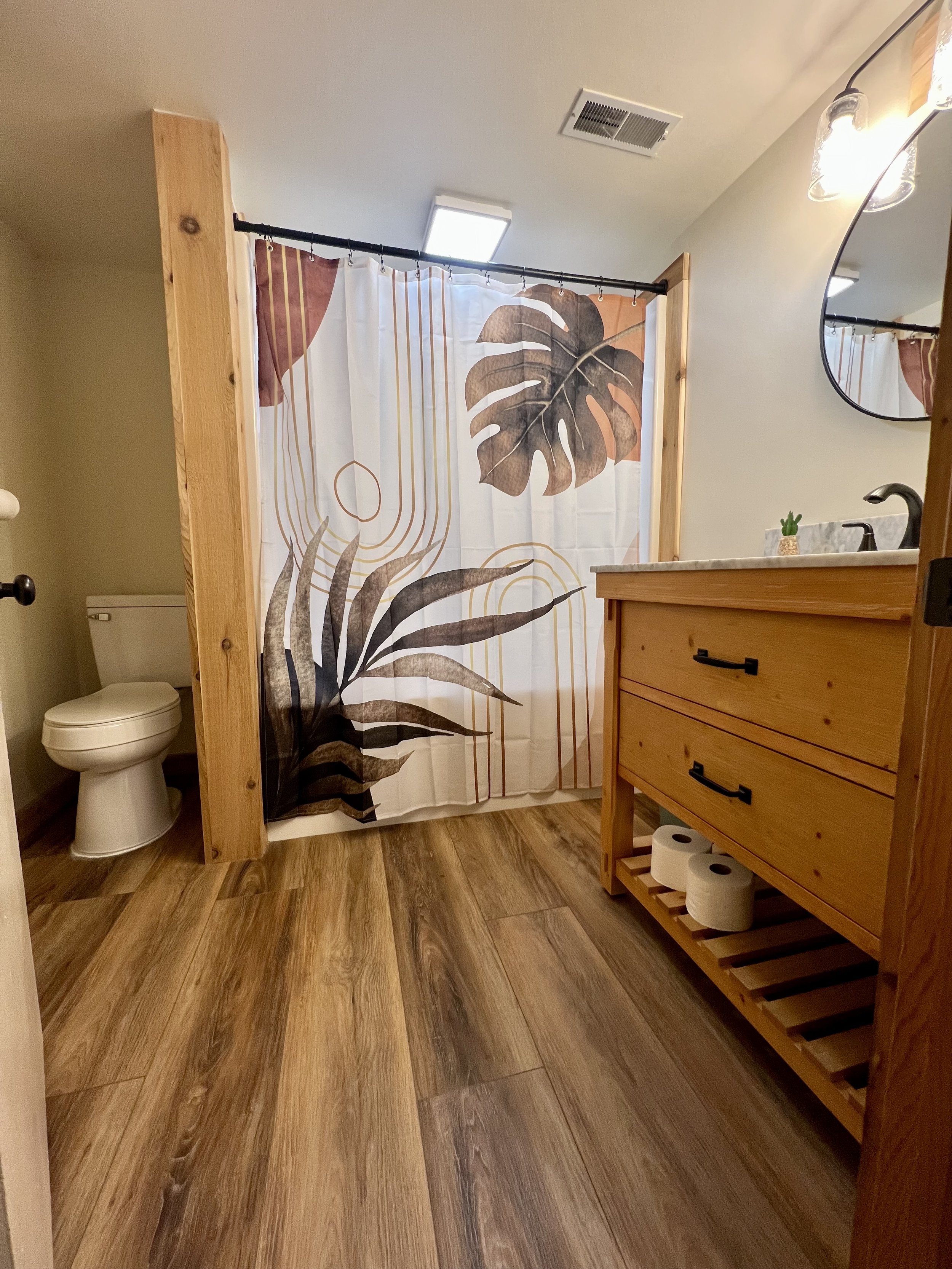 Bathroom with wooden accents, a shower curtain with large tropical leaf design, a toilet in the corner, a wooden dresser with rolls of toilet paper, a mirror, a small plant, and wood flooring.