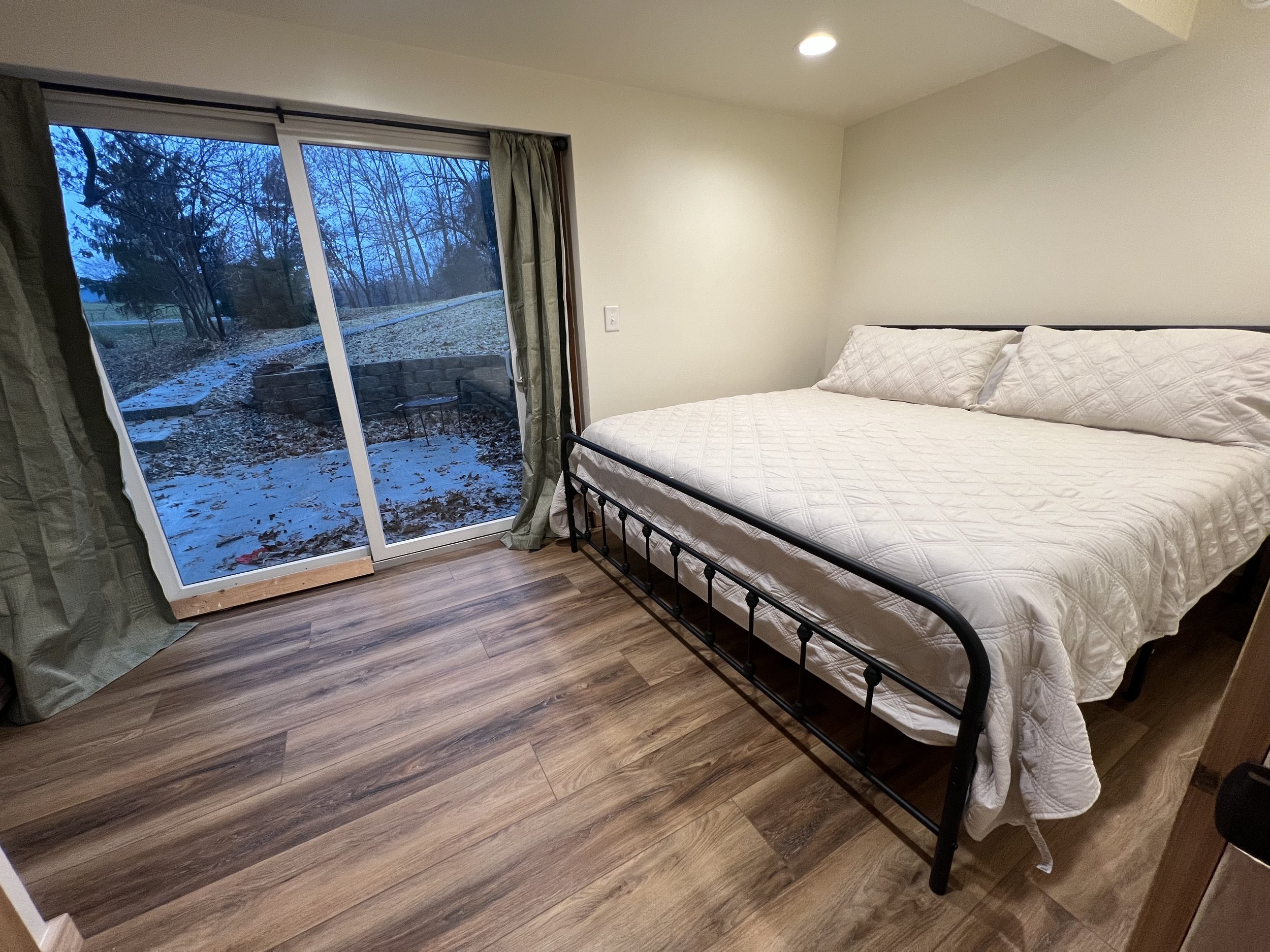 A bedroom with wooden flooring, a bed with white bedding, and a sliding glass door leading outside to a yard covered with fallen leaves. Outside are leafless trees and a hint of blue sky.