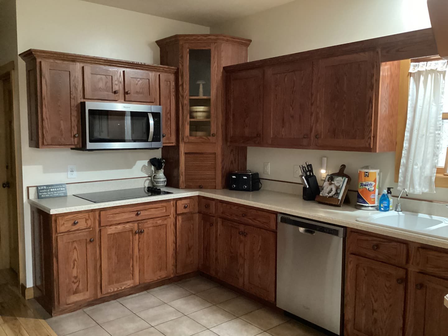 Kitchen with wooden cabinets, stainless steel microwave, countertop with small kitchen appliances, dish soap, paper towels, and a sink, window with white curtains.