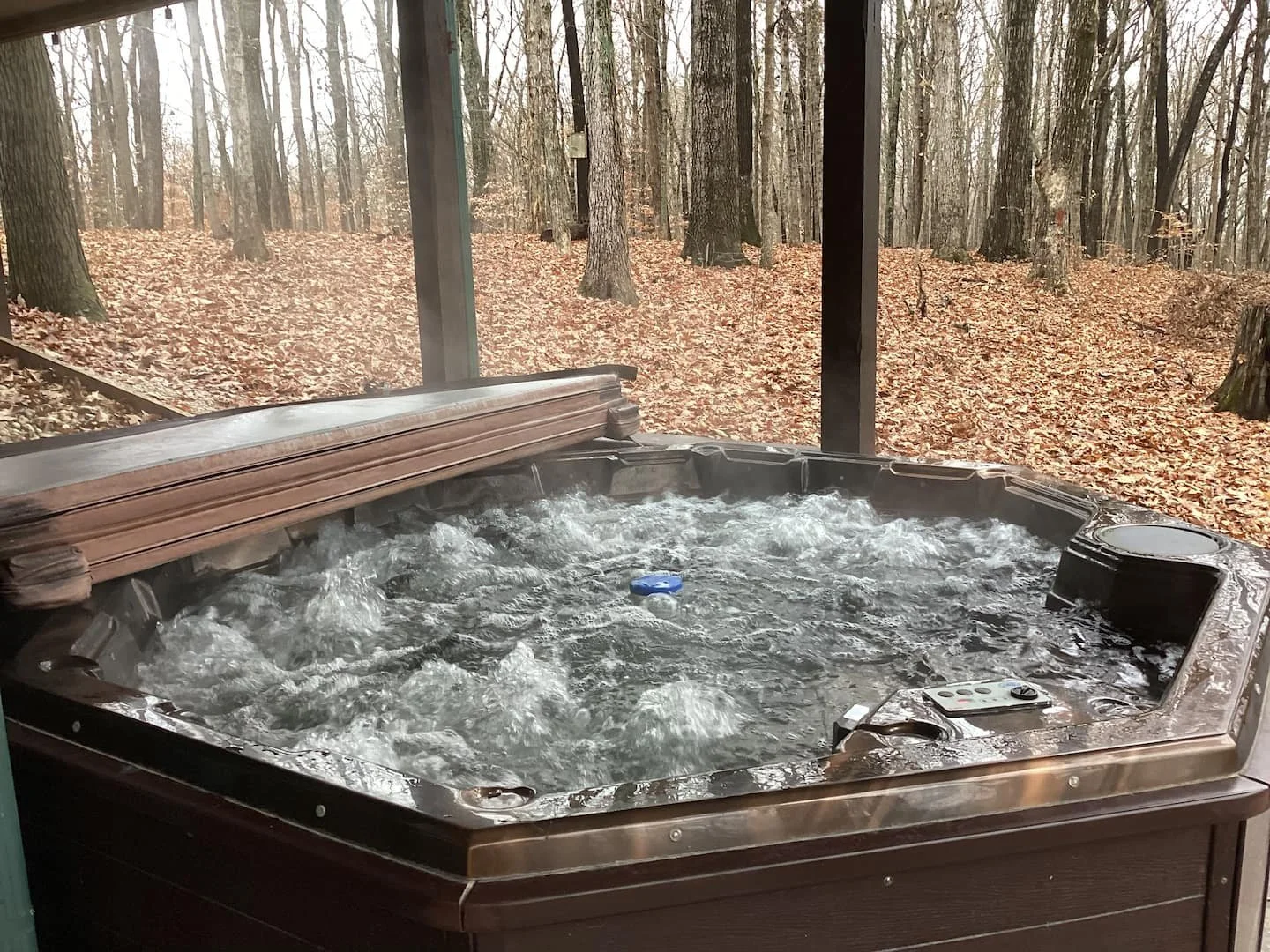 Hot tub with bubbling water on a porch overlooking a wooded area with fallen leaves.