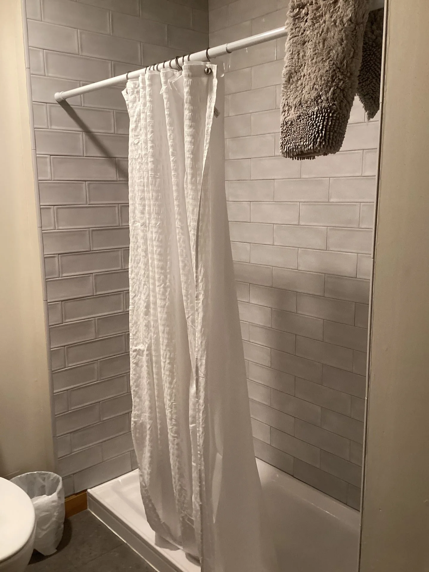 A bathroom shower area with a white curtain hanging from a rod, gray tiled walls, and a fluffy gray towel hanging on the wall. A small trash bin is visible at the bottom left corner.