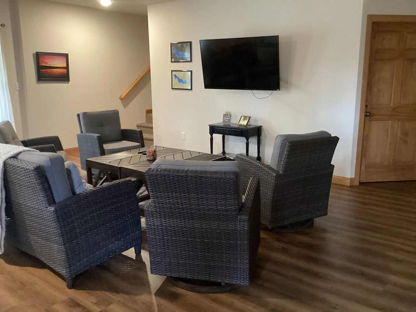 Living room with six dark wicker chairs around a rectangular wooden table, with a framed sunset picture on the wall, a staircase, a black TV mounted on wall, and a small table holding picture frames and a decorative item, with wood flooring and a woo