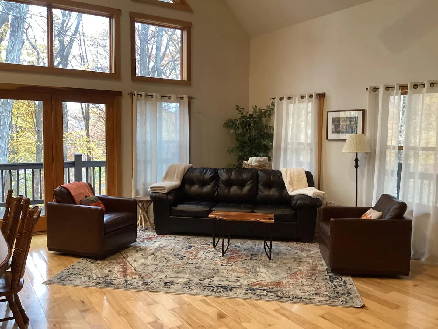 Living room with large windows showing trees outside, containing a black leather sofa, two matching armchairs, a wooden coffee table, a patterned area rug, a floor lamp, and curtains, with a potted plant in the corner.