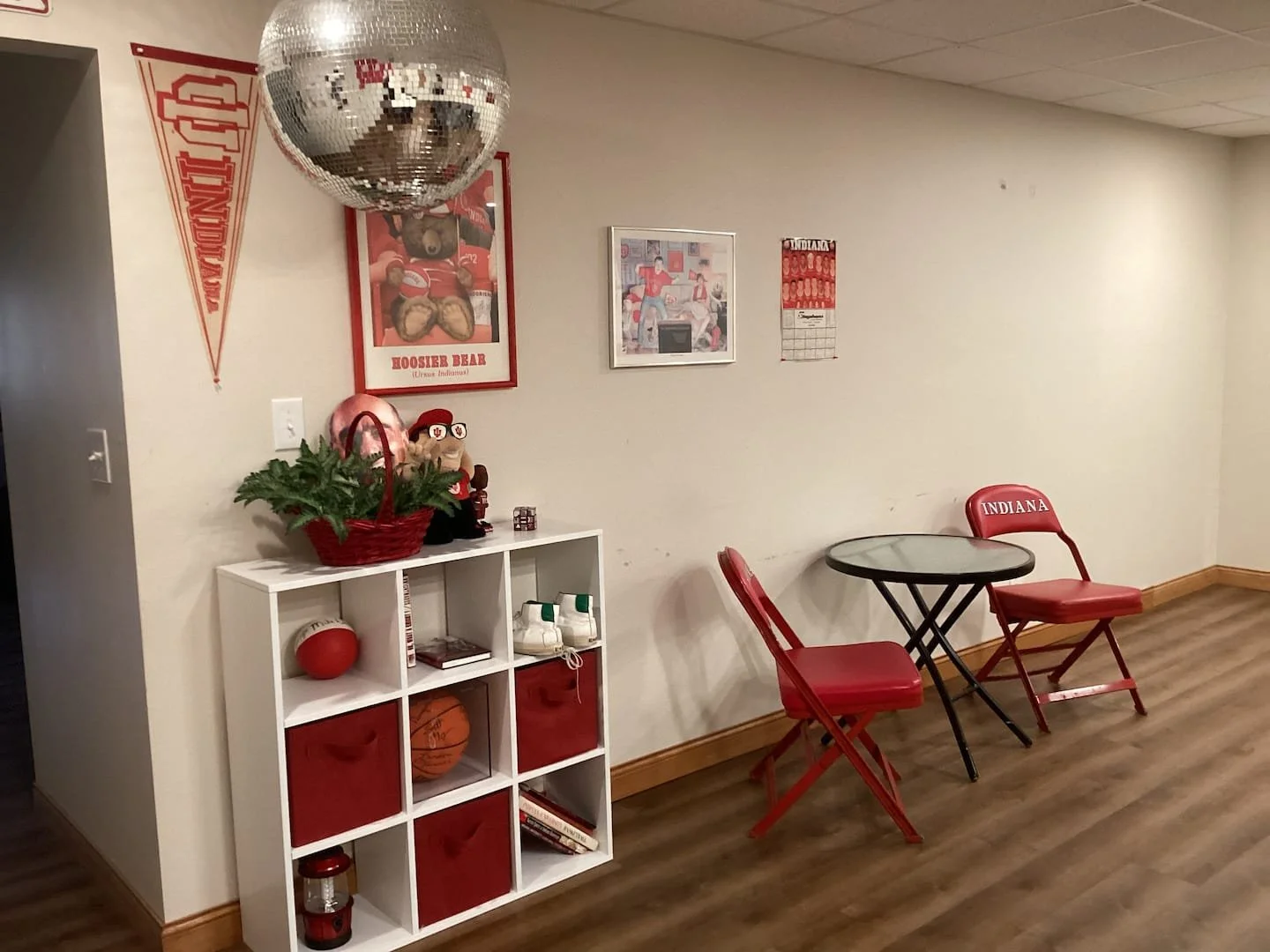 A corner of a room decorated in Indiana University sports theme, with Red chairs, a small round table, framed sports posters, a white cubby shelf with sports memorabilia, a disco ball, and Indiana University flags.