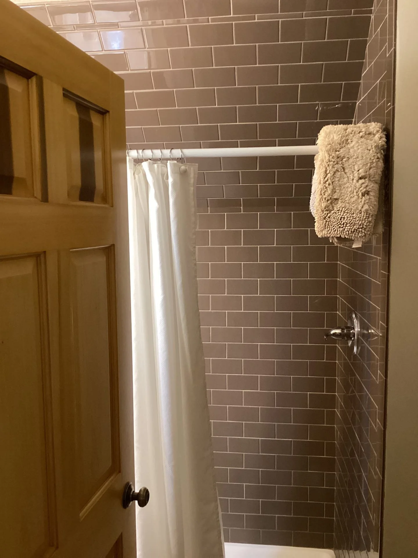A bathroom with brown subway tiles on the wall, a white shower rod, a white shower curtain, a beige fluffy towel on a hook, and a silver faucet.