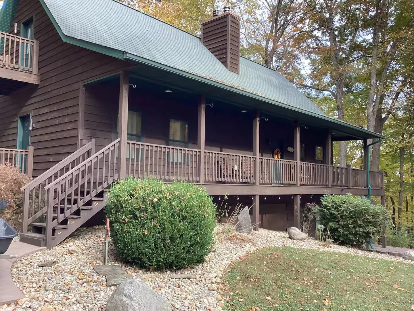 Back of a brown wooden house with a large covered porch, stairs leading up, surrounded by trees and landscaped rocks and bushes.