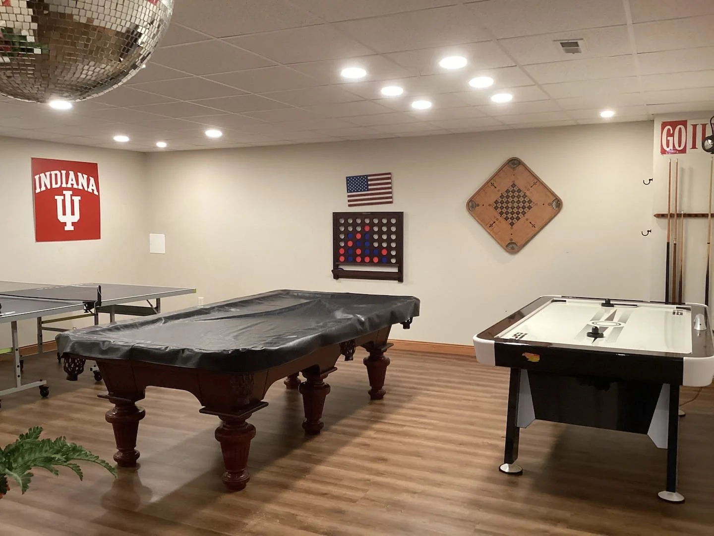 A game room with a pool table covered with plastic, an air hockey table, a ping pong table, and wall decorations including an Indiana University banner, an American flag, a connect four game, and a vintage shuffleboard and cue sticks.