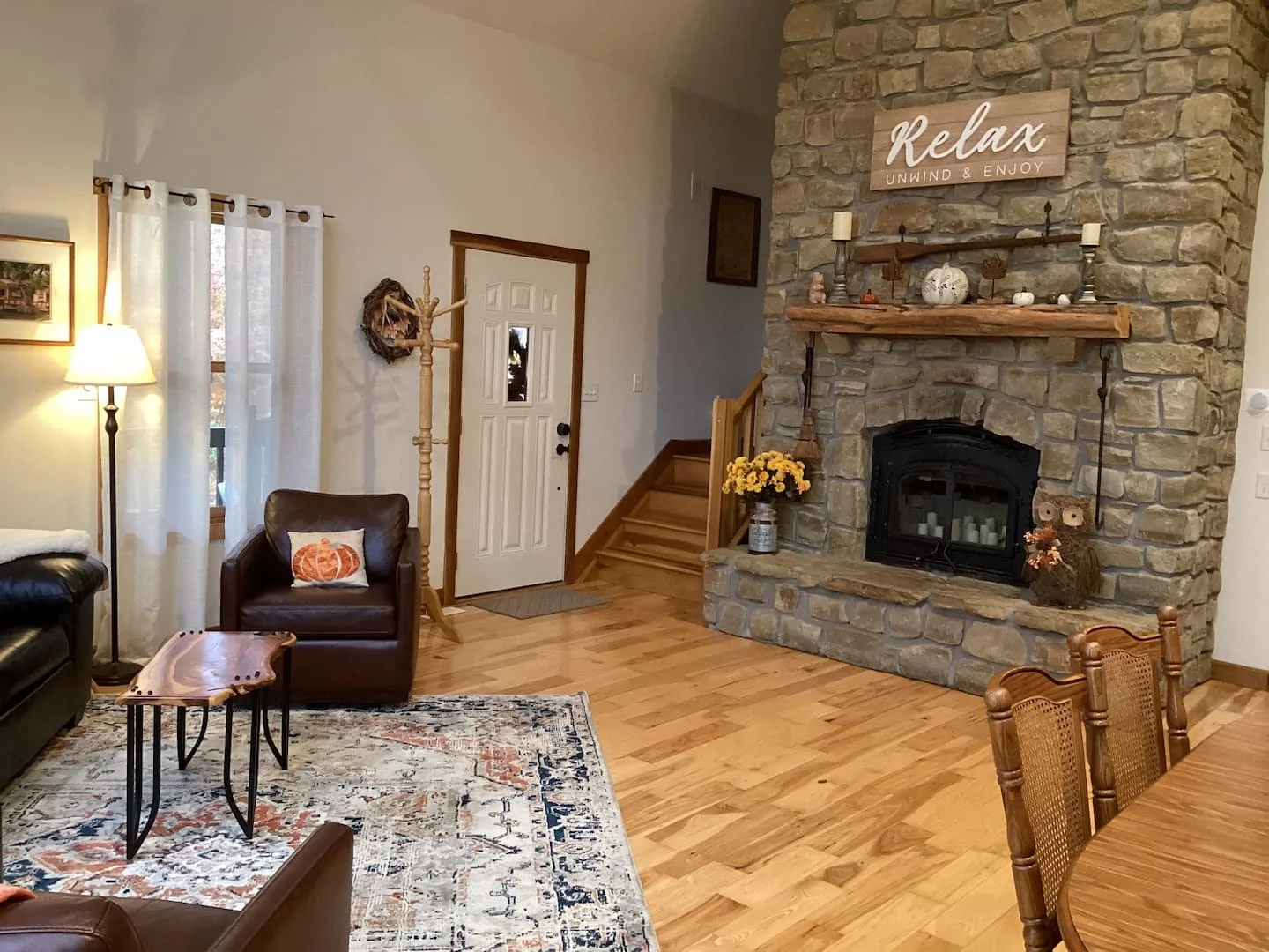 Living room with a stone fireplace, wooden mantel decorated with candles and small pumpkins, a sign that says "Relax Unwind & Enjoy," a brown leather armchair with a pumpkin pillow, a wooden side table, a area rug, curtains, a lamp, and wooden stairs