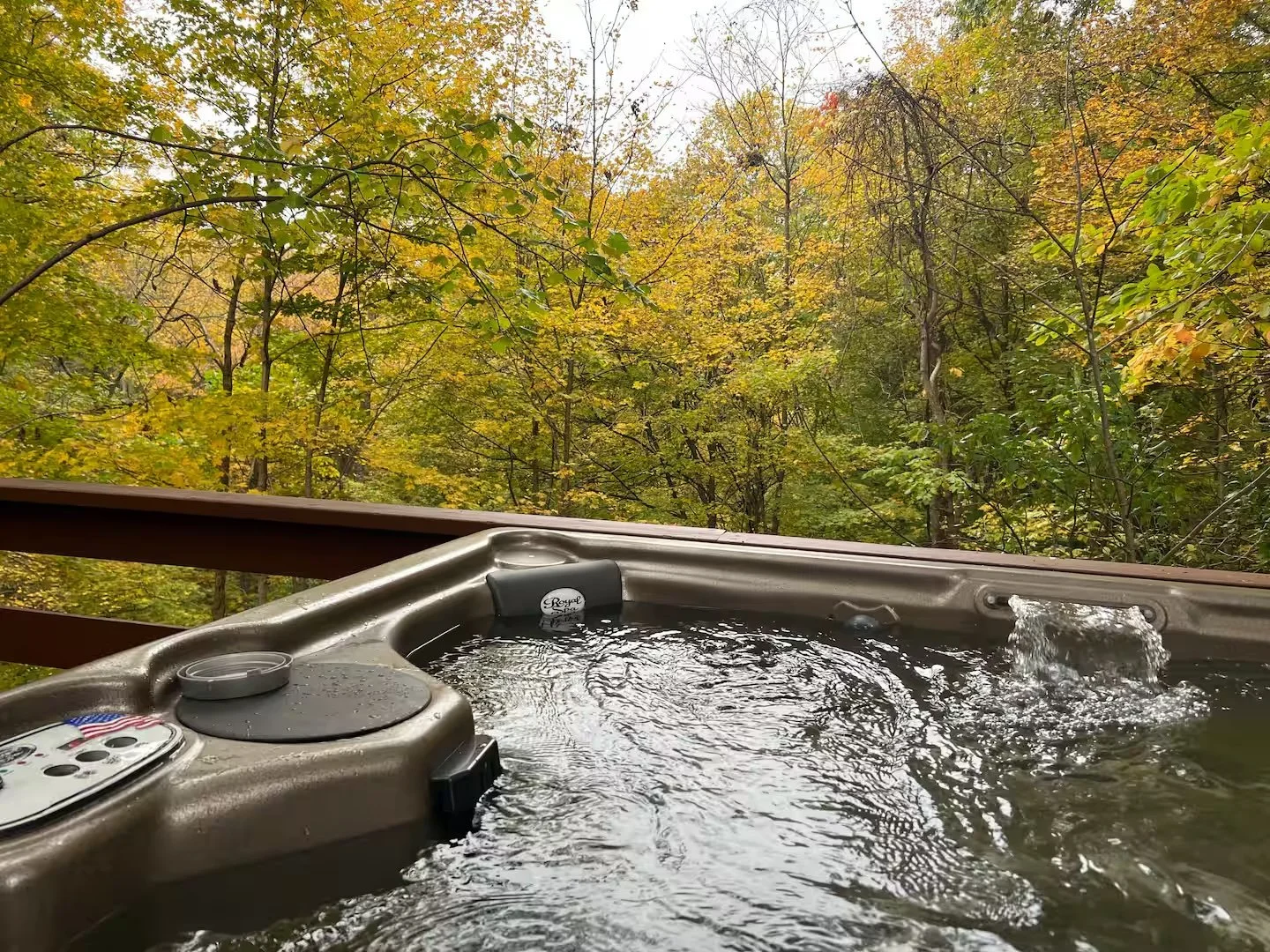 Hot tub with water jets on a wooden deck surrounded by autumn foliage with green, yellow, and orange leaves.