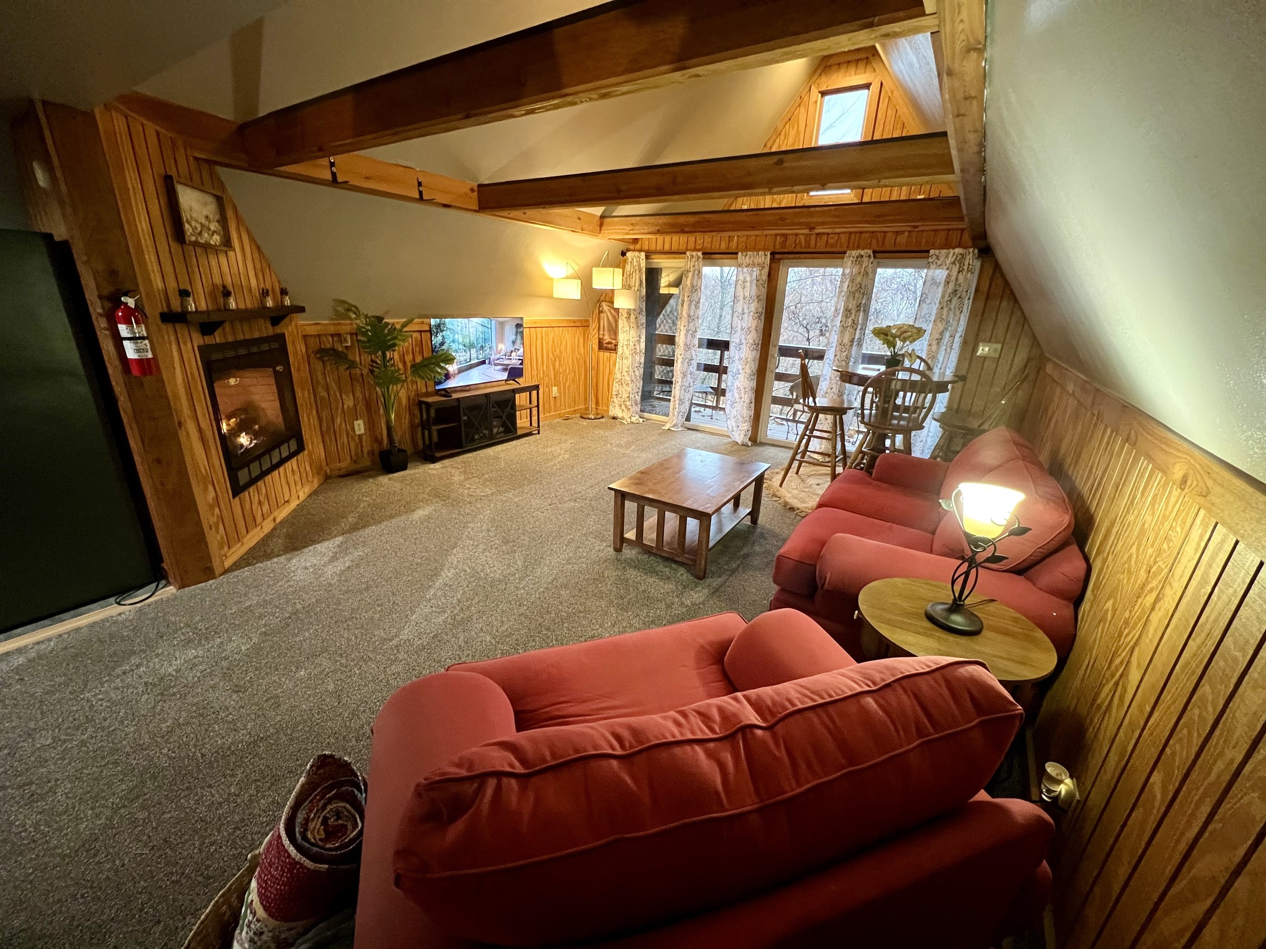 Living room with wood-paneled walls and vaulted ceiling, featuring a red sofa, a small wooden coffee table, a fireplace, a potted plant, and a balcony with sliding glass doors covered by floral curtains. There are lamps providing warm lighting.