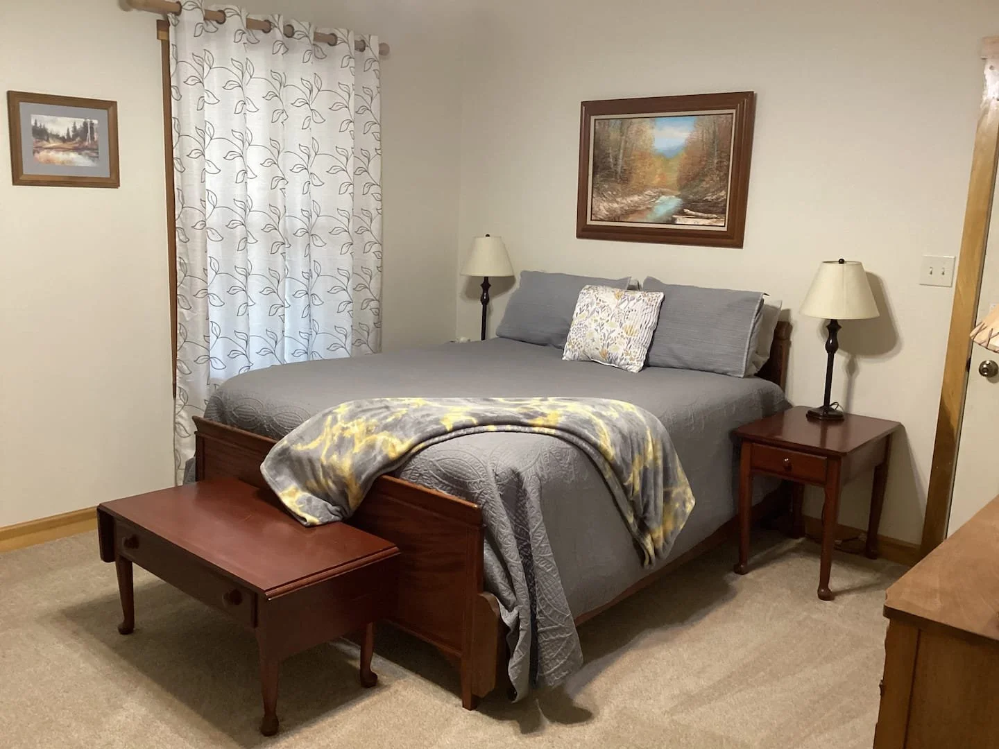 A bedroom with a bed covered in grey bedding, a throw blanket at the foot, and a decorative pillow. There are two bedside tables with lamps, and wall art above the bed. A window with patterned curtains is on the left wall.