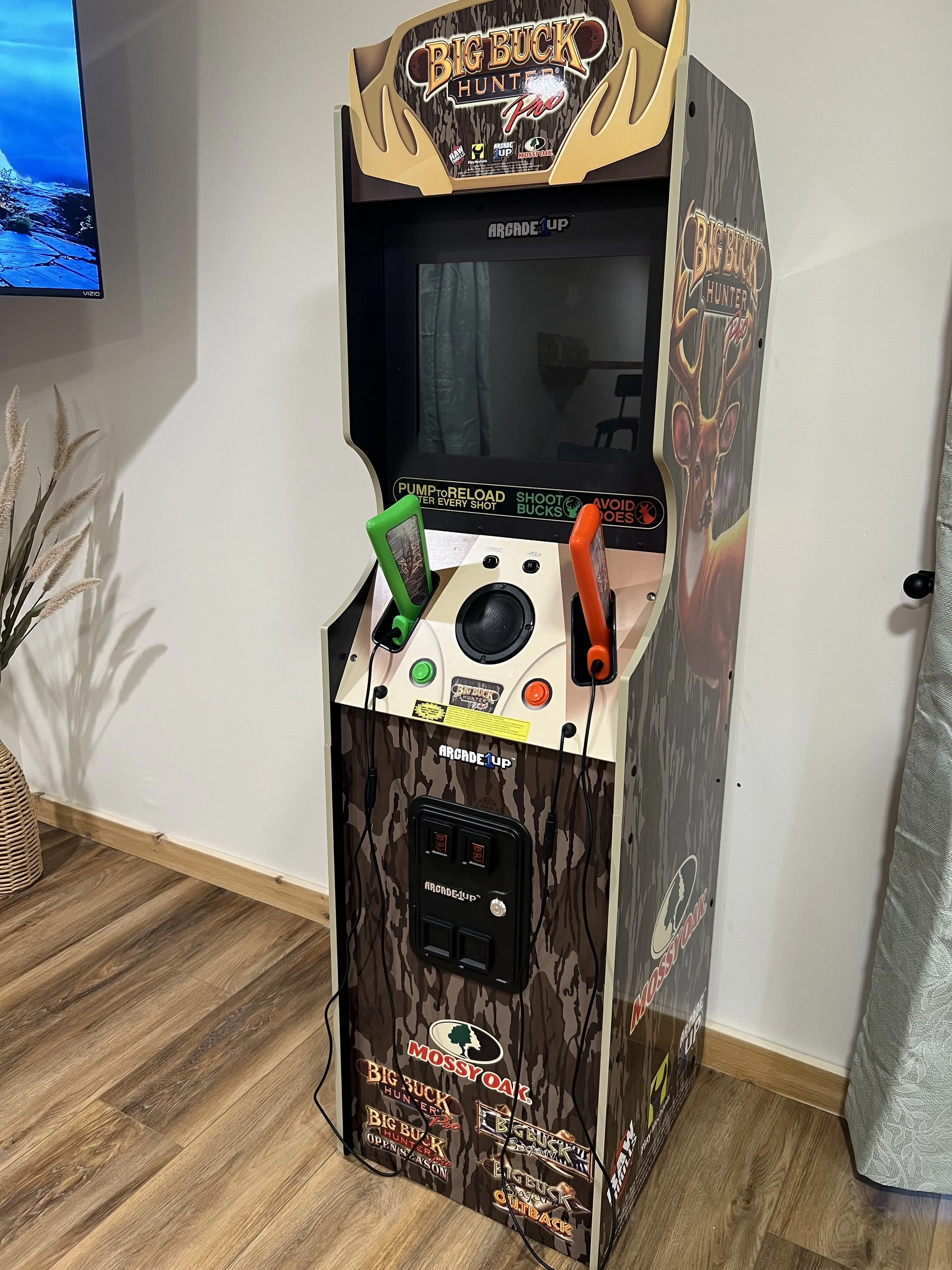 An arcade game machine titled 'Big Buck Hunter Pro' with a camouflage pattern on the side and front, featuring a deer graphic. The machine has two controller guns, one green and one orange, attached to it. The screen is off, and there are instruction