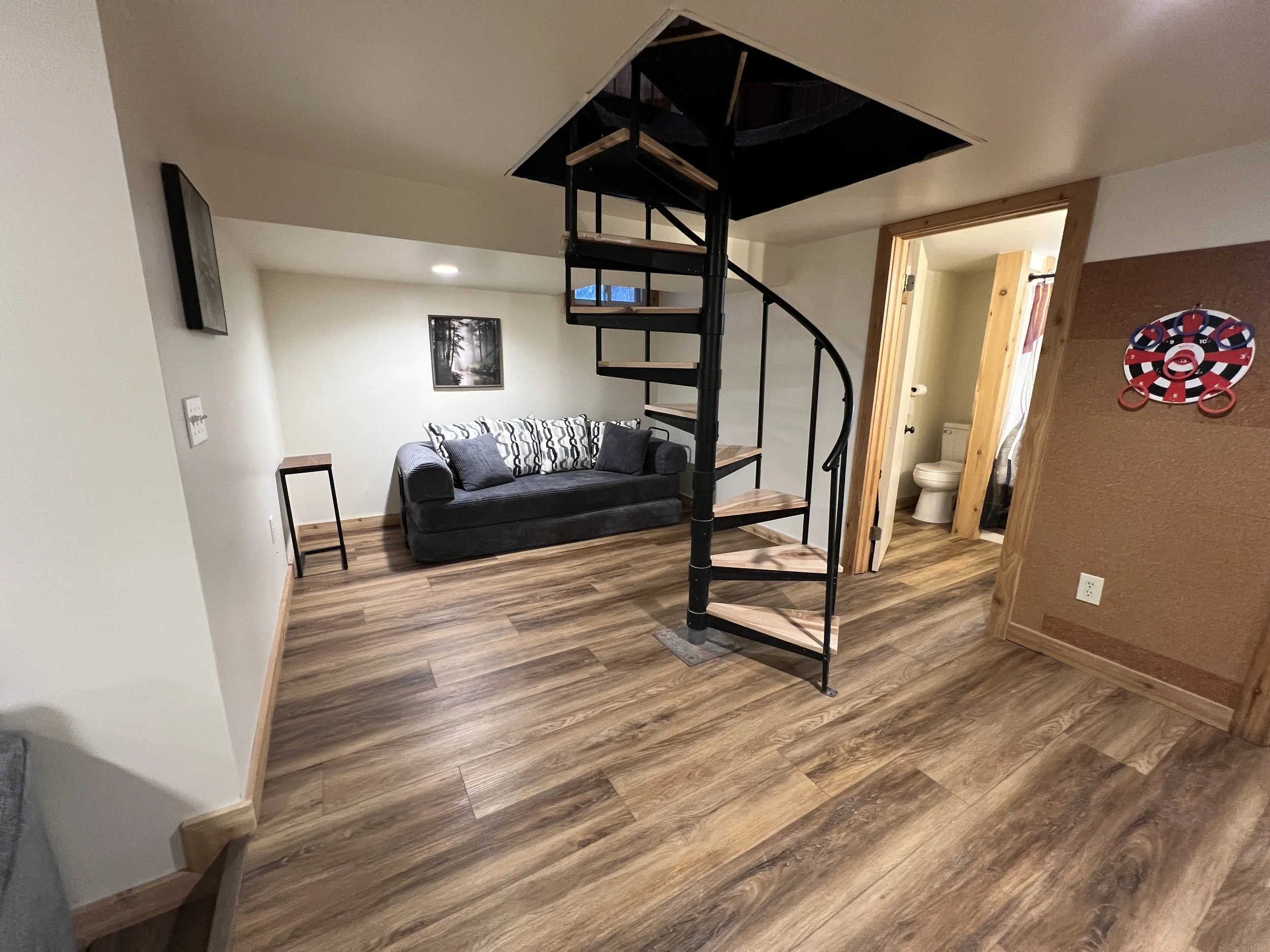 Interior of a living room with a black sofa, a small table, and a picture of a forest on the wall. A spiral staircase with wooden steps and black metal railing leads upwards. The room has wood flooring and trim, and an open doorway reveals a bathroom