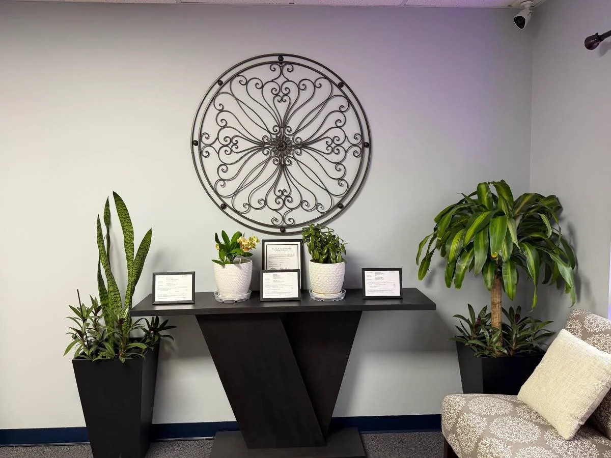 A decorative wall with a large circular metal art piece, a black table with three framed documents, and potted plants. There is a upholstered chair with a cushion on the right.