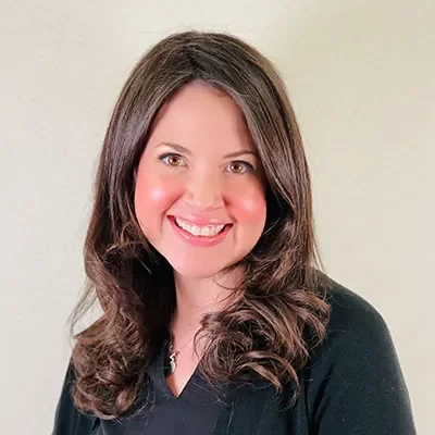 A woman with long, wavy brown hair and fair skin, smiling and wearing a black top, standing against a neutral background.
