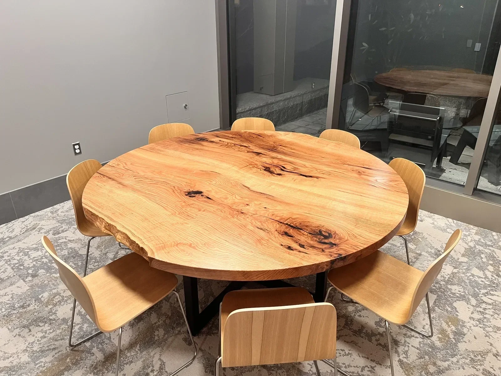 Round Red Oak table with a natural, live edge finish, surrounded by seven light wooden chairs with metal legs, inside a university classroom.