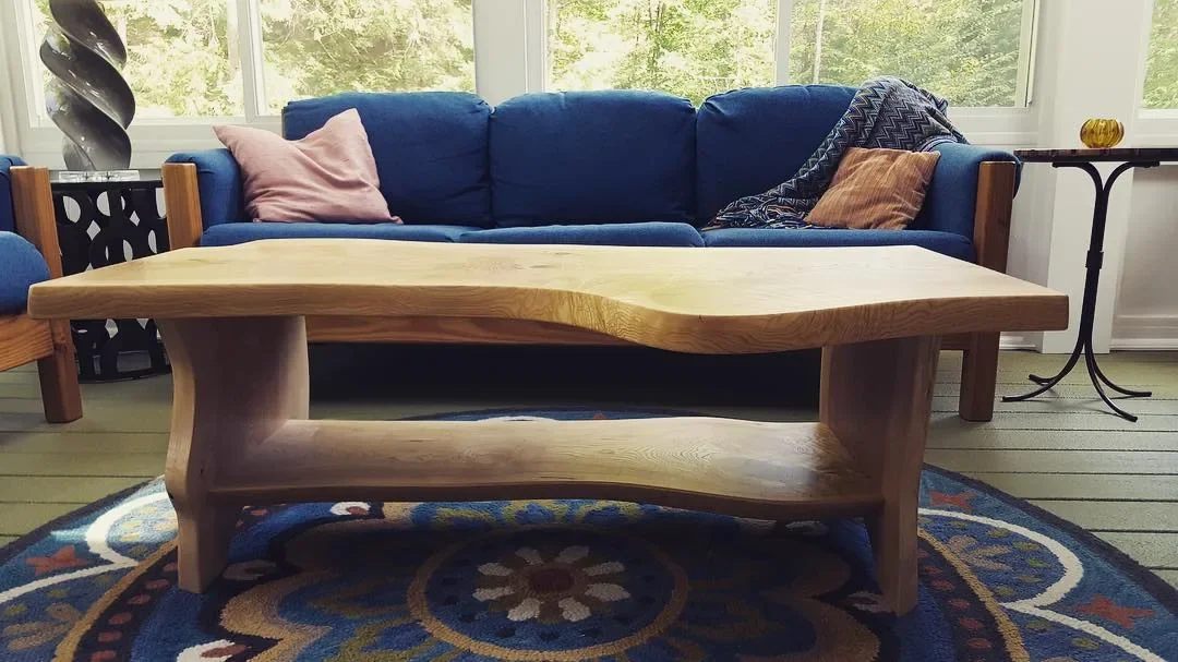 Custom live edge coffee table with lower shelf and natural wood legs, installed in a sunroom alongside a blue sofa