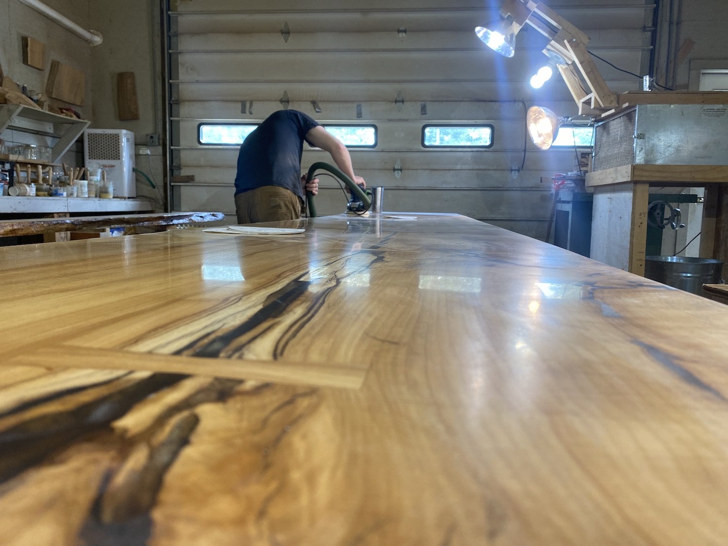 Lead artisan in vtg workshop sanding or finishing a wooden table using a handheld power tool, with workbenches and lighting fixtures in the background.